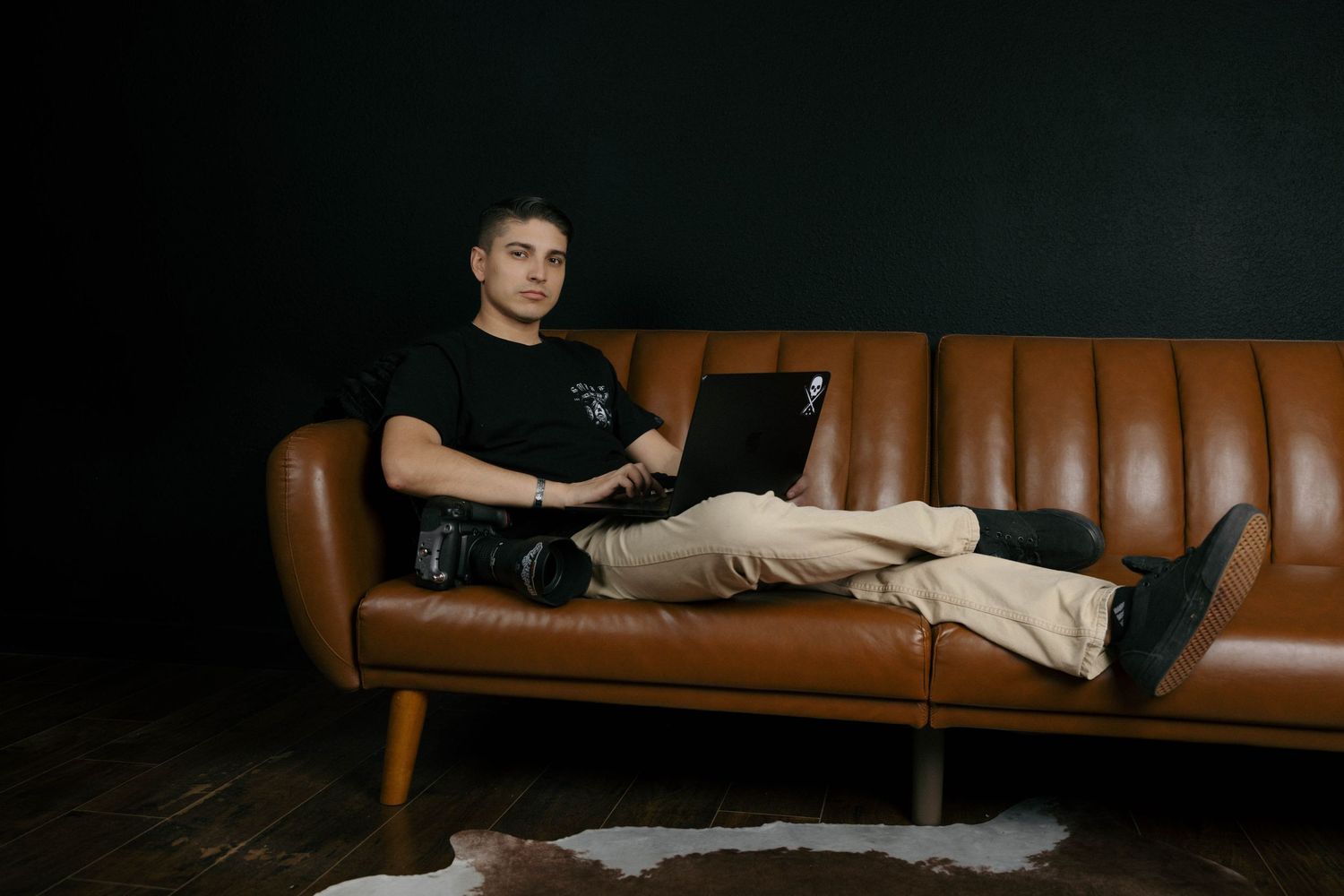Man relaxing on leather sofa with laptop and camera in studio.