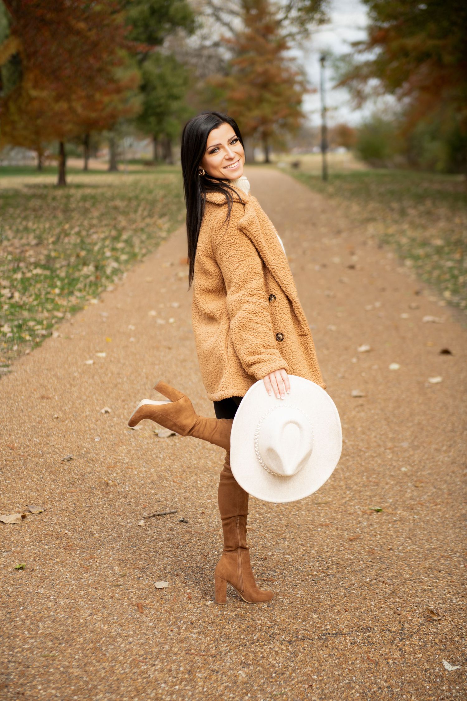 Smiling brunette woman in camel coat holding white hat while walking in autumn park.