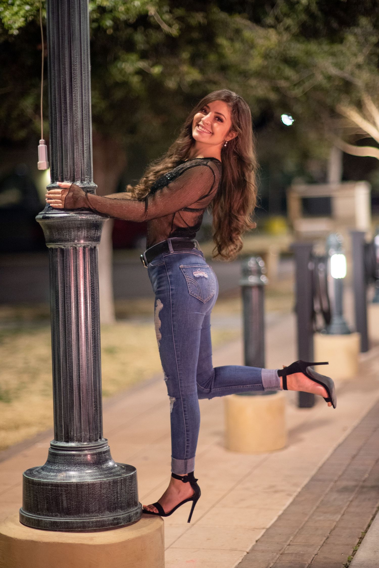 Smiling woman in sheer black top and jeans leaning on decorative lamp post at night.