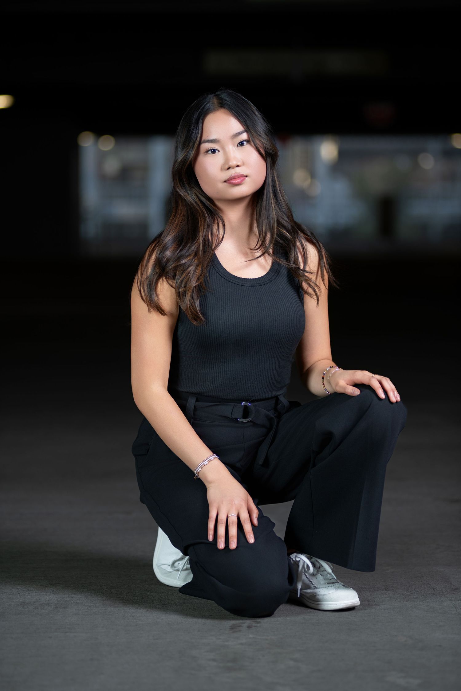 Young Asian woman in black tank top and wide-leg pants posing in a crouching position in a dark parking garage.