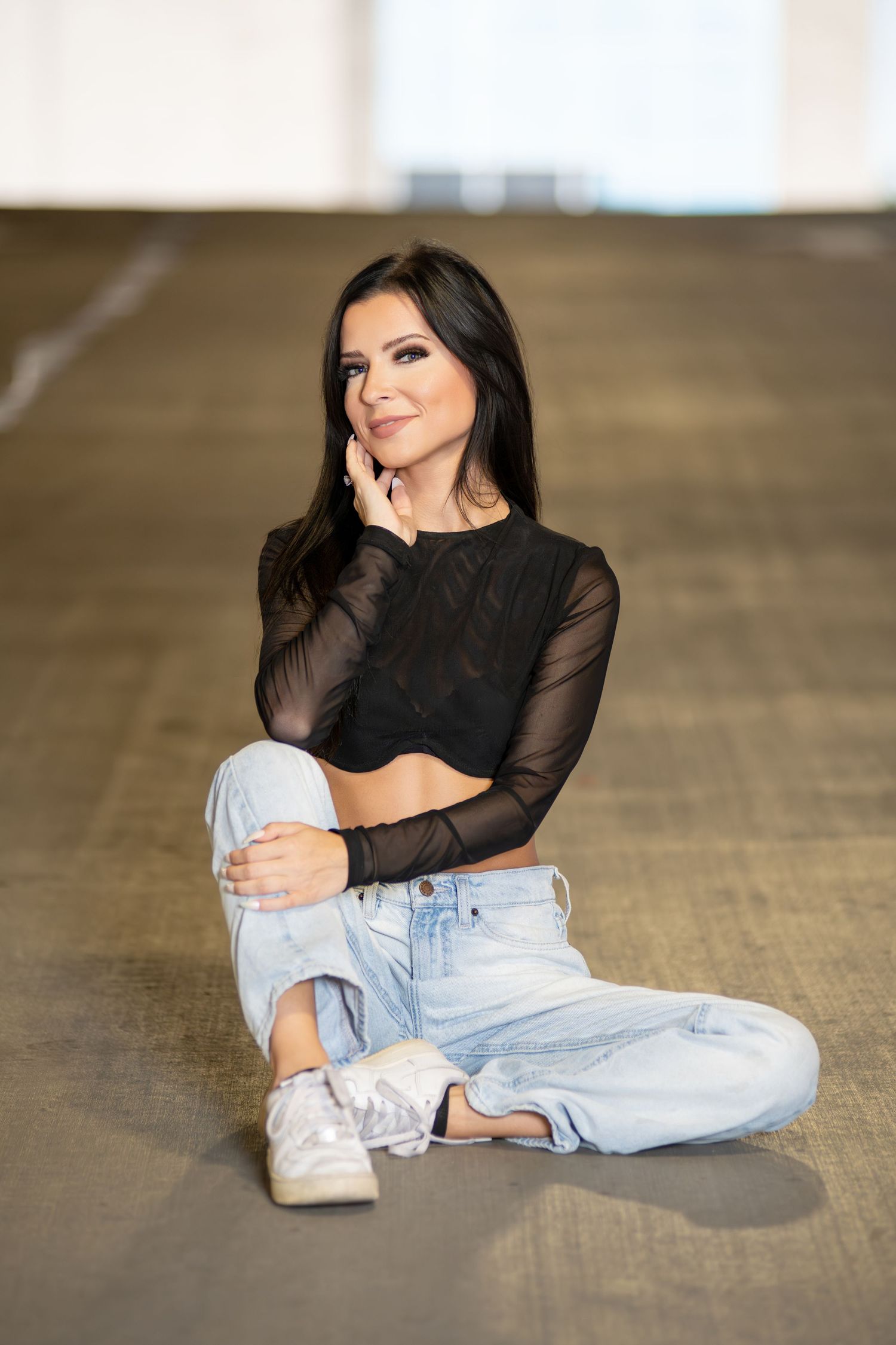 Brunette woman in black mesh crop top and light jeans sitting on floor in bright space.