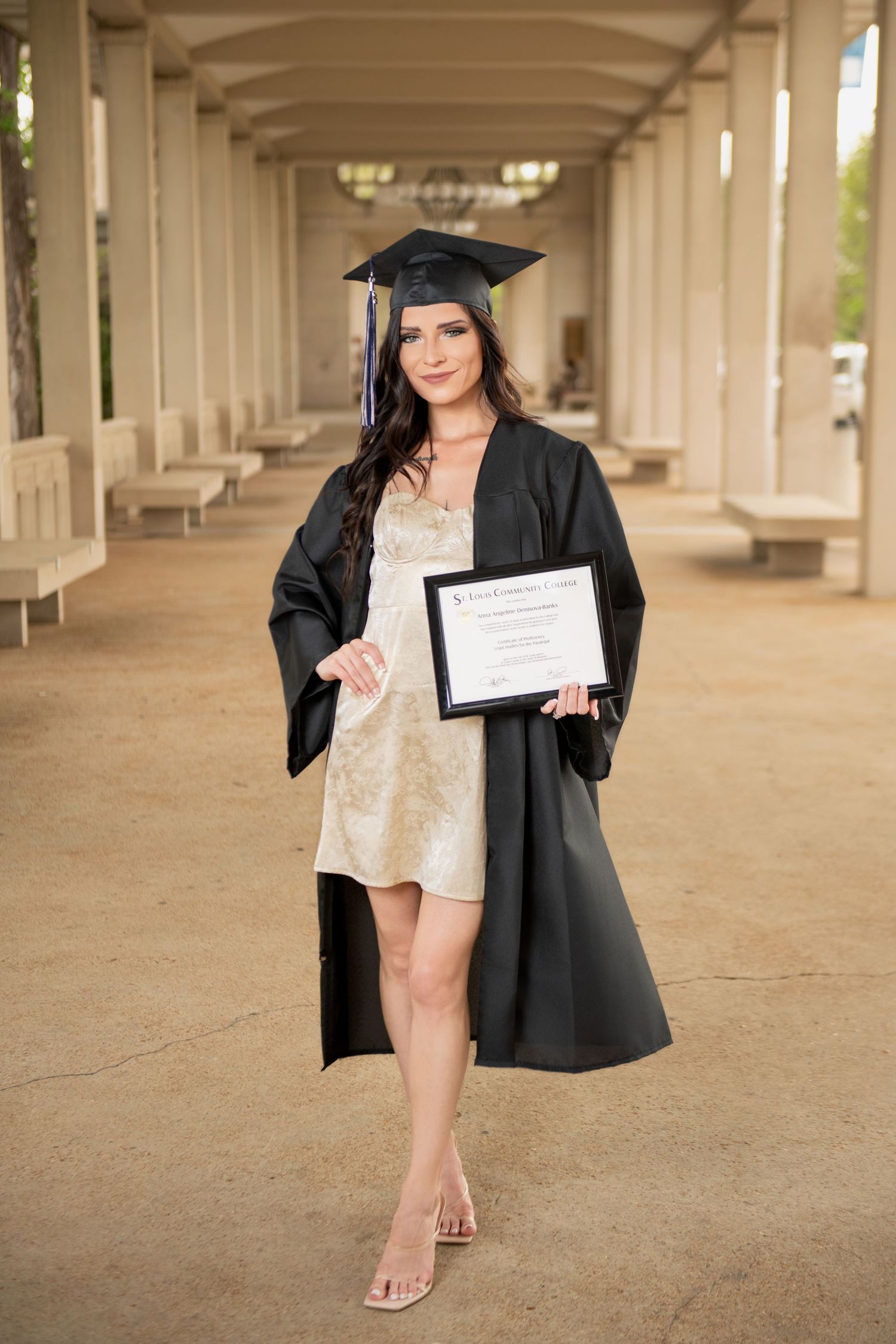 Female graduate in cap and gown proudly displaying diploma under an outdoor colonnade.