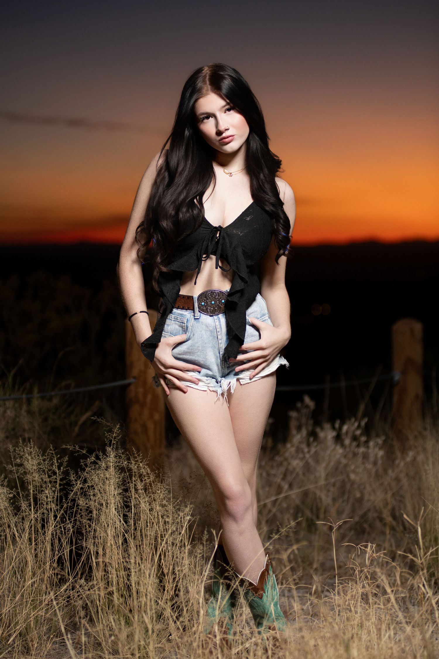 Young woman in black top and denim shorts poses in field against vivid sunset sky.