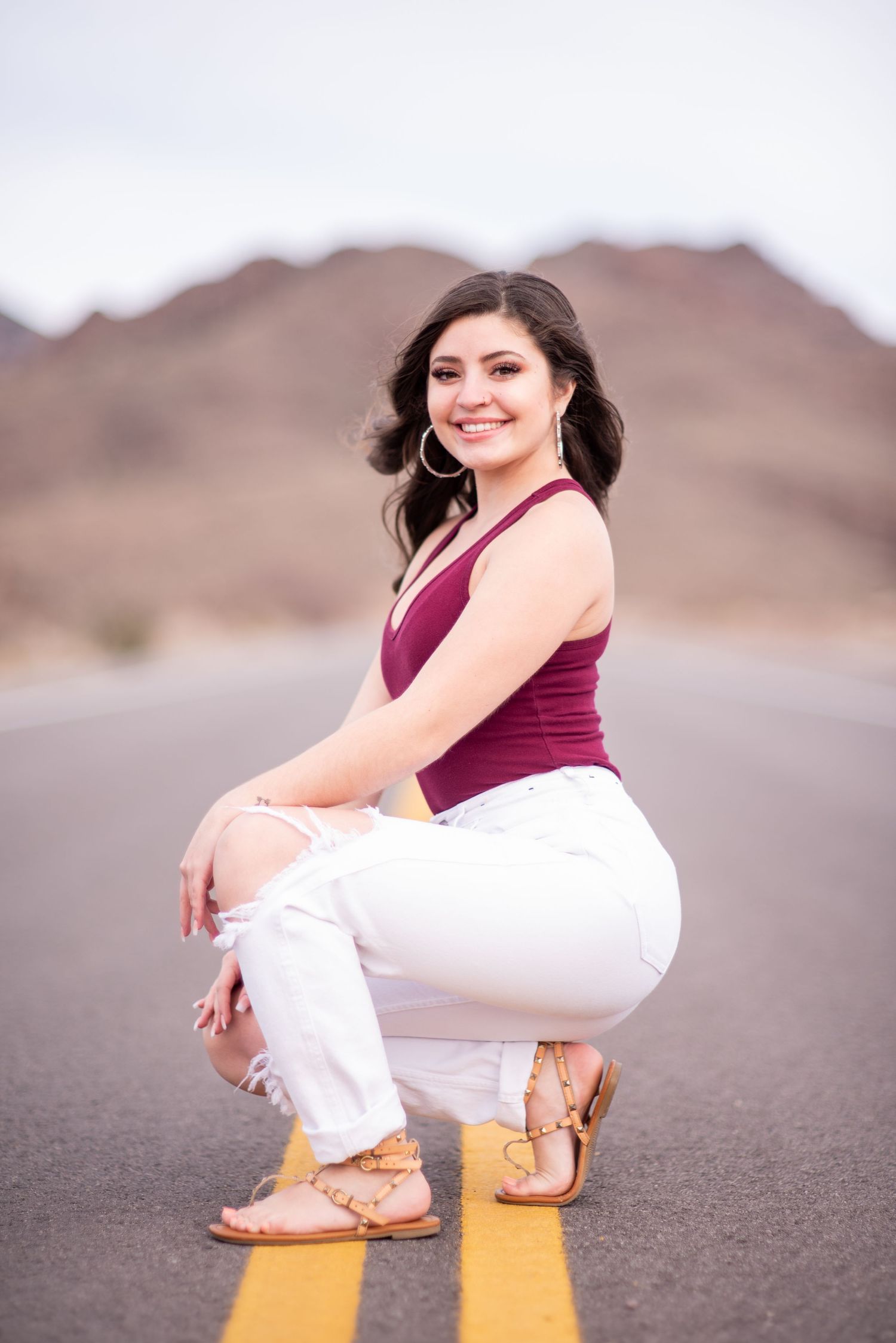 Smiling woman in burgundy top and white ripped jeans posing on desert road.