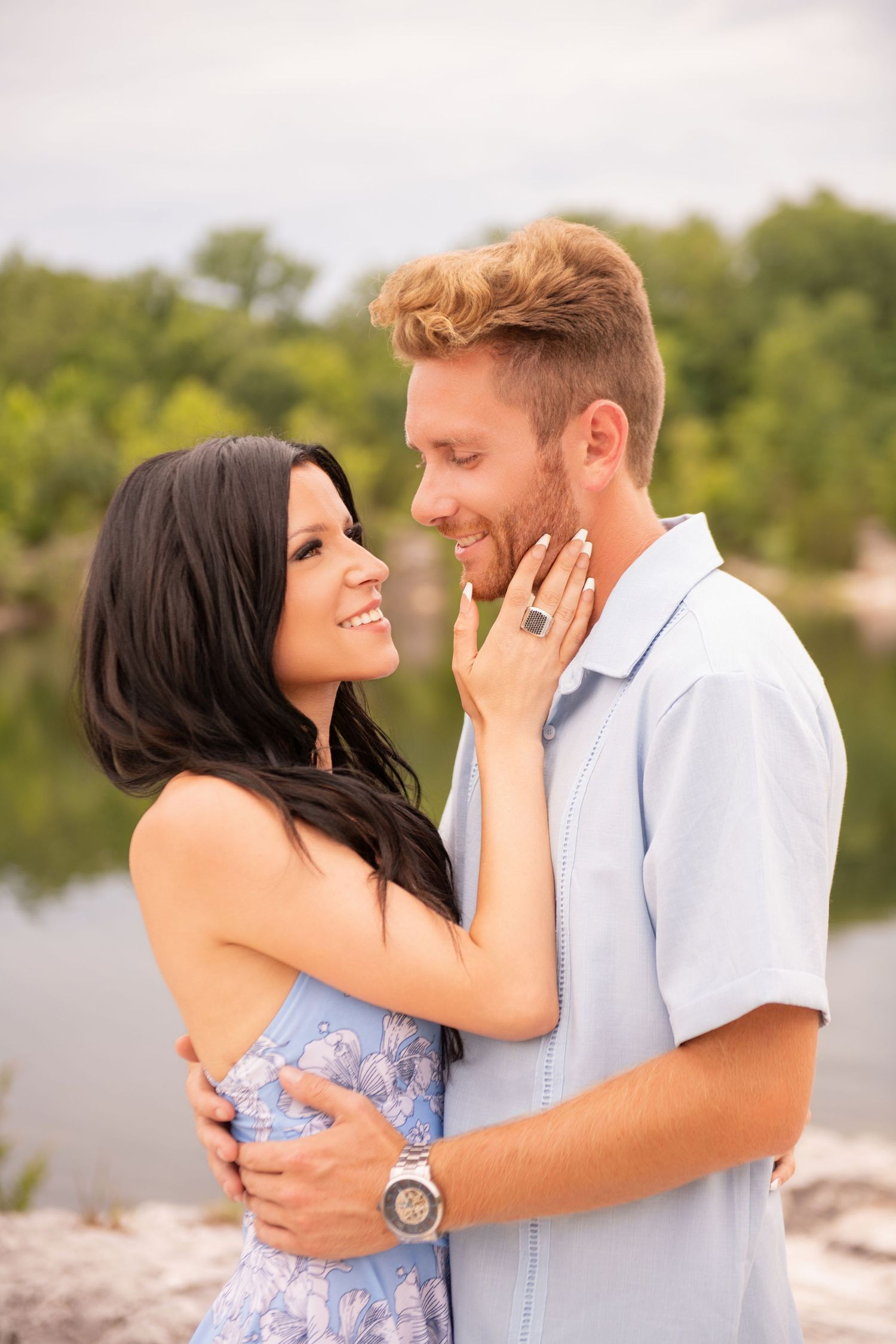 Smiling couple embracing outdoors, woman touching man's face, showing engagement ring, with lake in background.