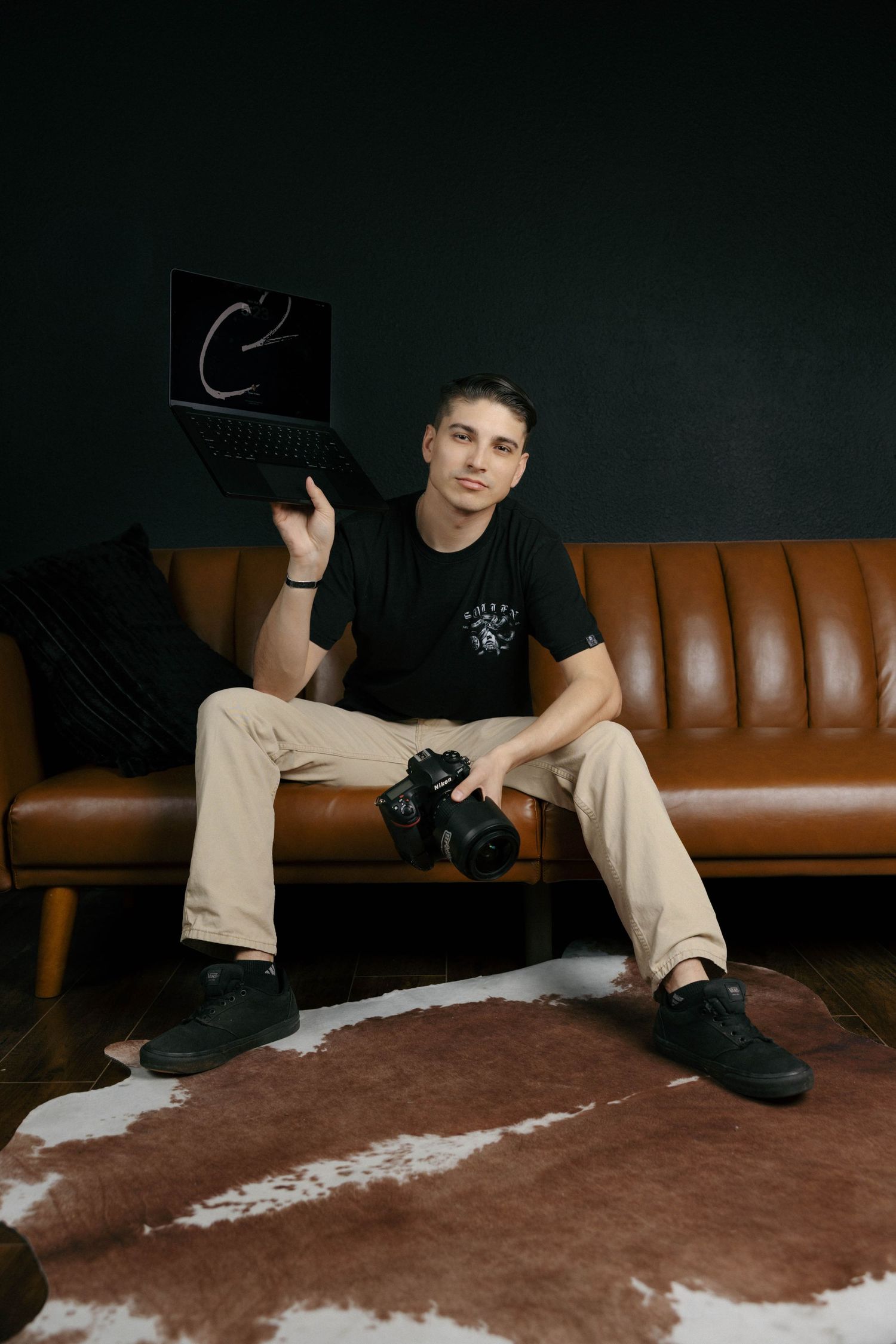 Young male photographer sitting on brown leather sofa holding laptop and camera in dark green studio.
