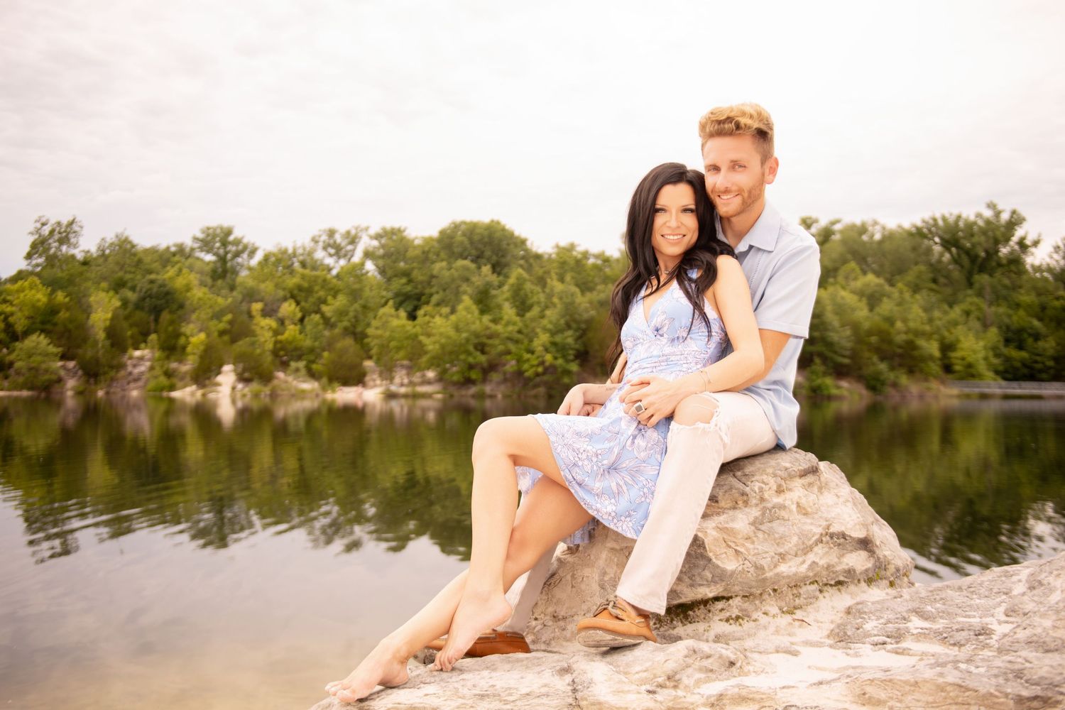 Romantic couple sitting on rocks by a lake, woman leaning back into man's arms, surrounded by lush green trees.