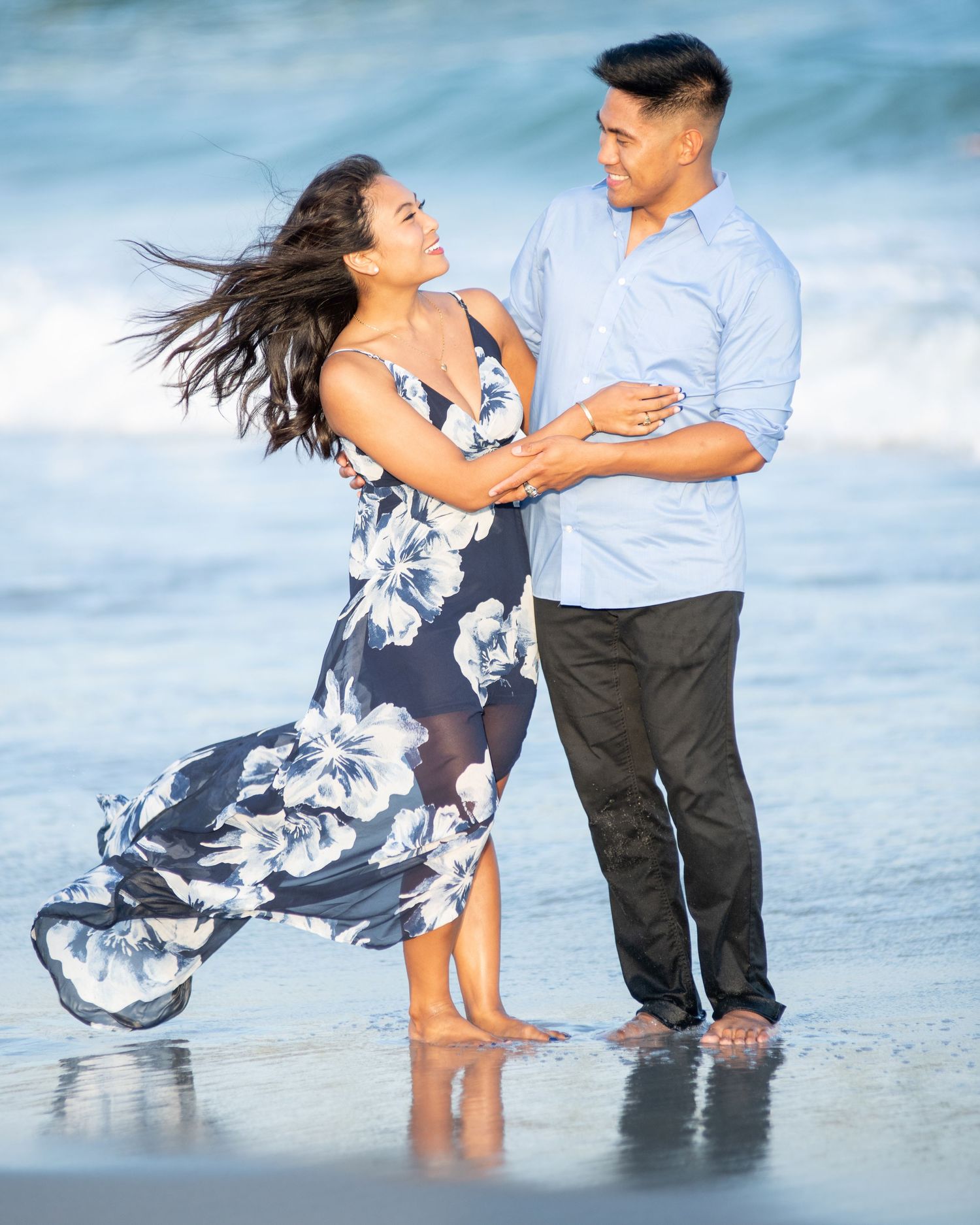 Couple dancing joyfully on a beach with ocean waves, woman in floral maxi dress.