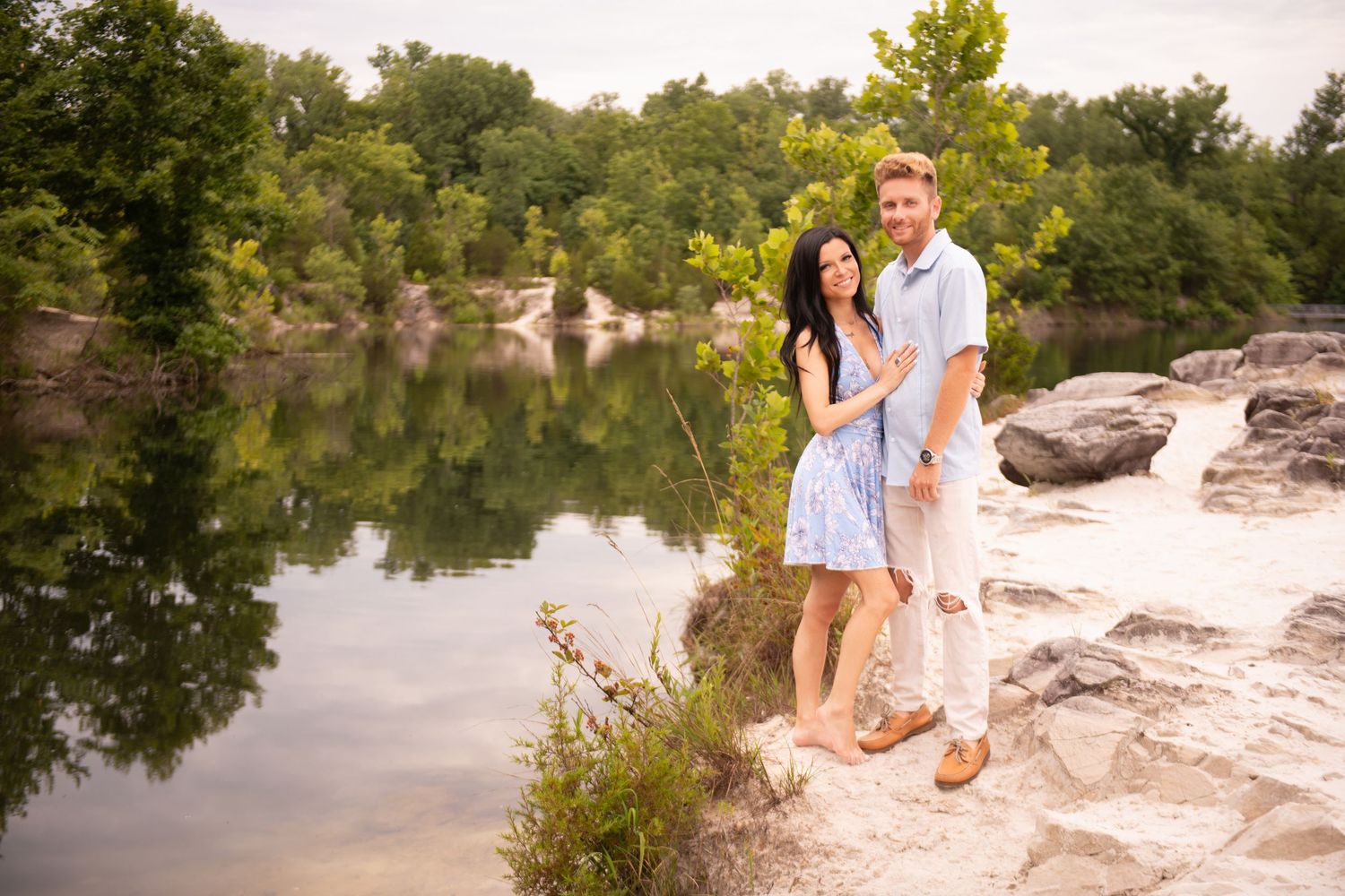 Engaged couple posing together on rocky lakeside, woman in blue dress, man in light blue shirt, green trees behind.