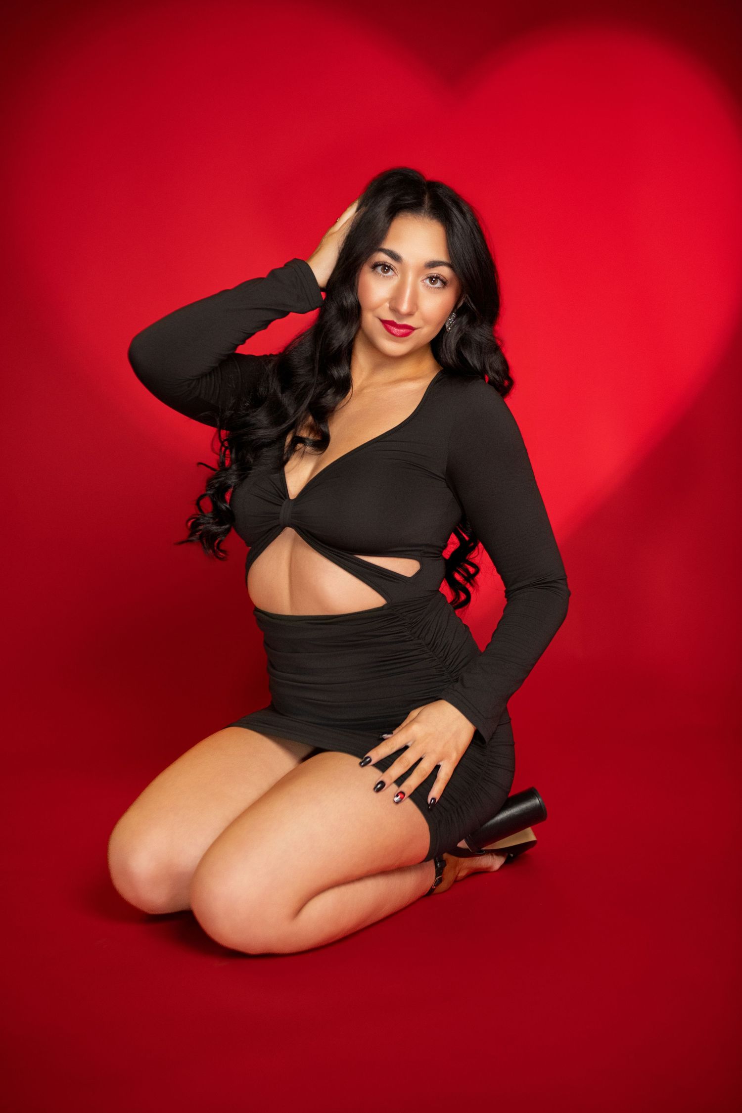 Glamorous woman in black cutout dress posing in front of a red heart background.