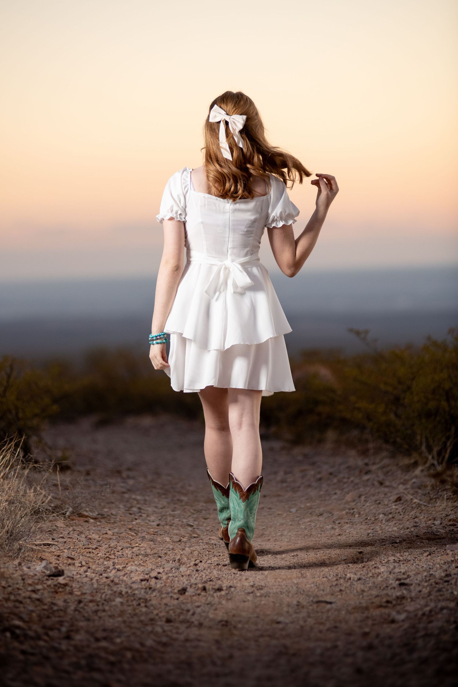 Young woman in white ruffled dress and turquoise cowboy boots walking down a dirt path at sunset.
