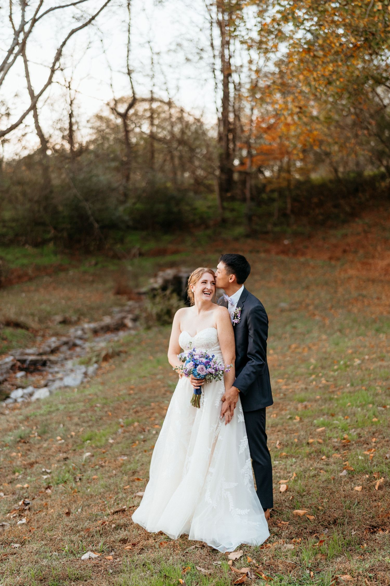 A romantic outdoor wedding couple walking through a scenic autumn forest setting with fallen leaves and a gentle stream nearby.