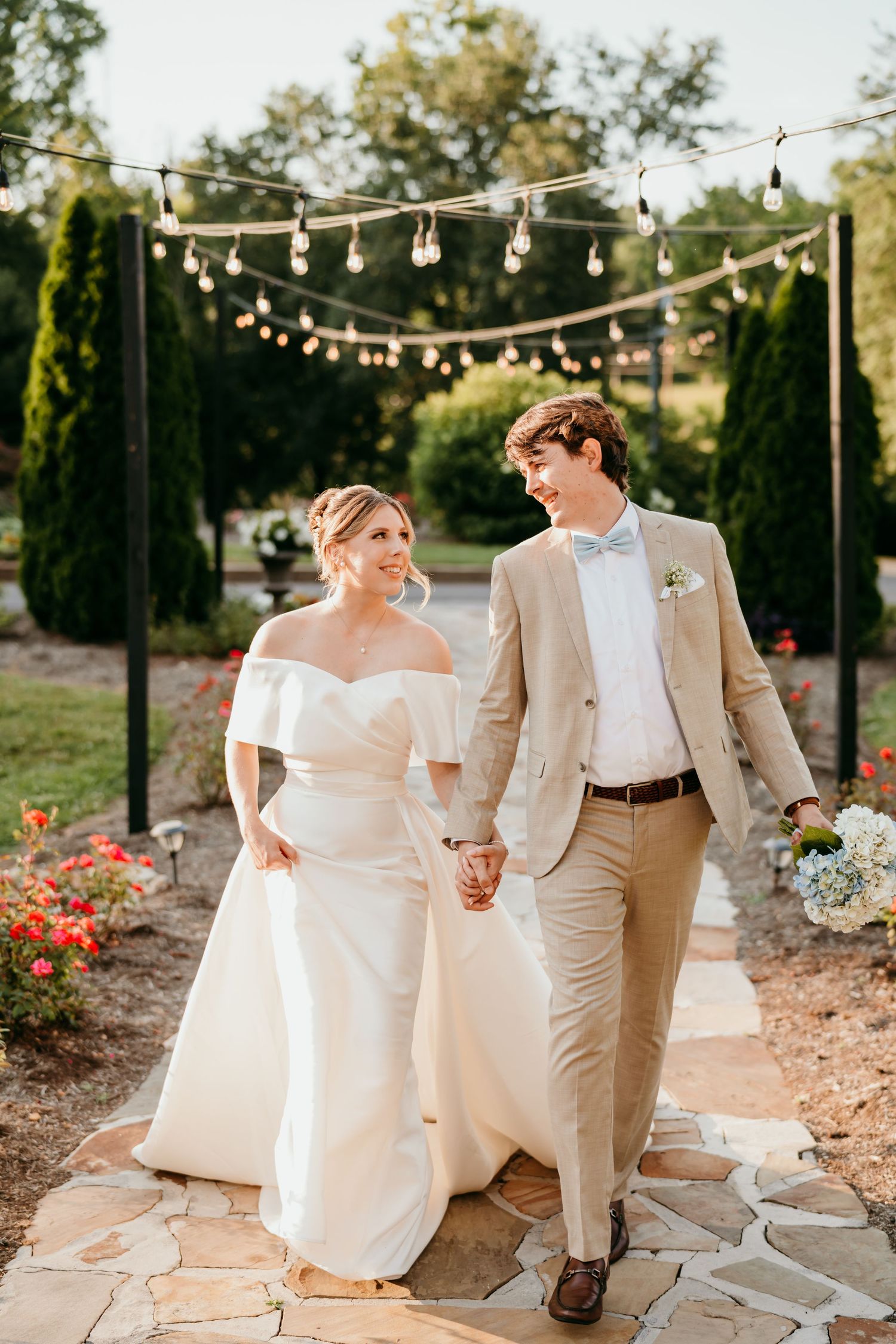 A couple in wedding attire sits on stone steps under twinkling string lights in a garden setting at golden hour.