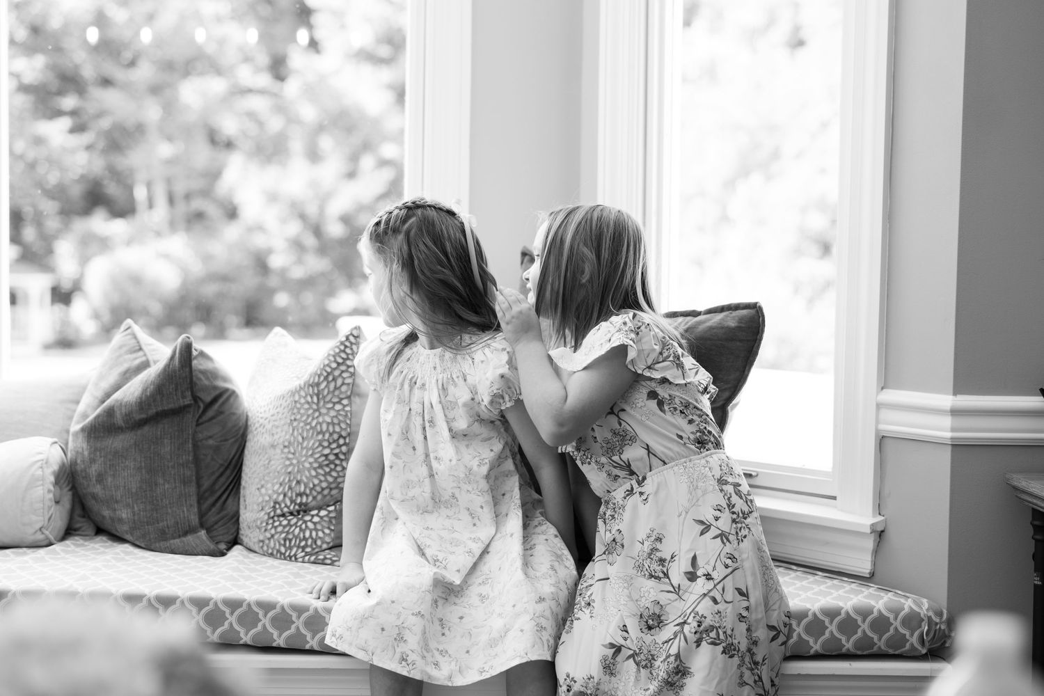Two young children in dresses looking out a sunny window together.