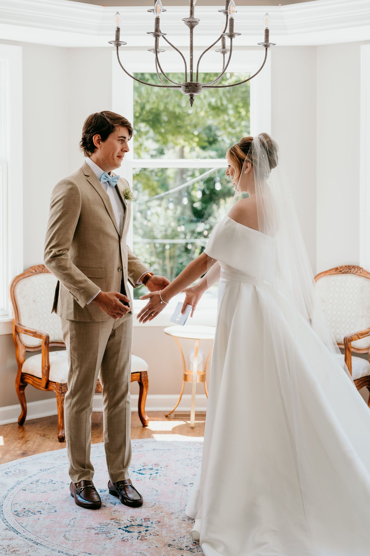 Wedding first look moment captured in a bright sunlit room with a tan suited groom and bride in traditional white dress.
