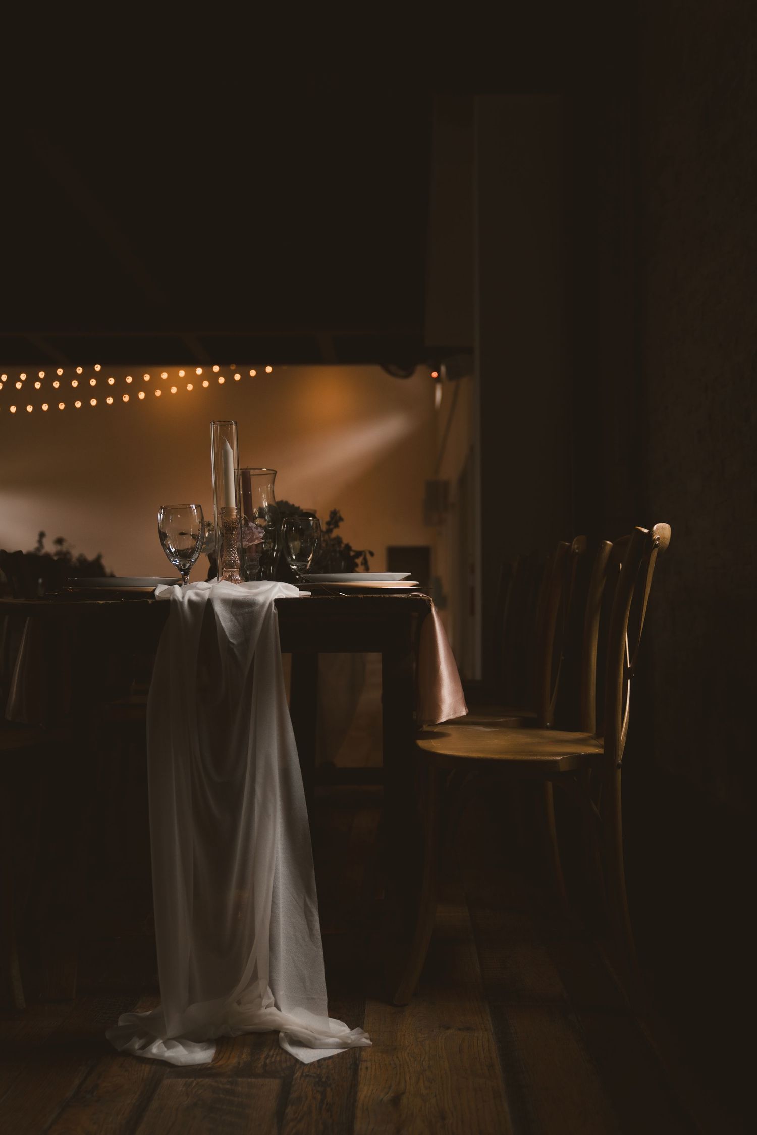 Moody wedding table setup with white tablecloth and string lights creating romantic ambiance in dark room.