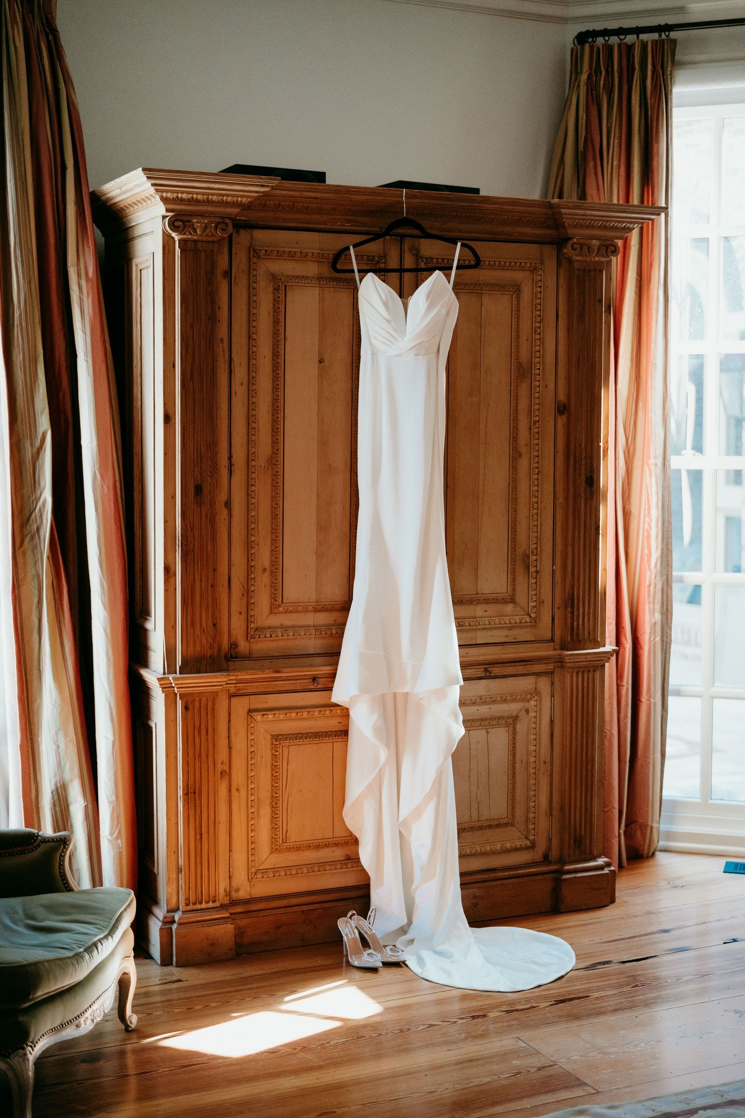 A sleek white wedding dress with spaghetti straps hangs on a wooden hanger in front of an antique armoire with golden curtains Nashville TN