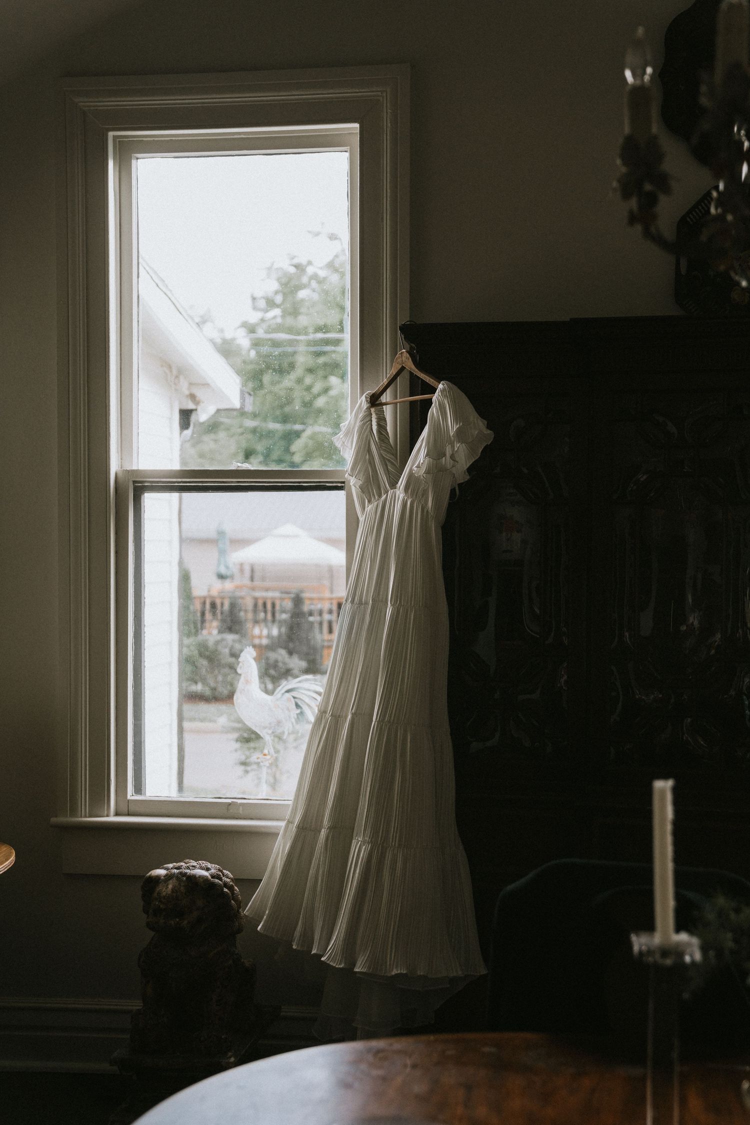 A white wedding dress hangs delicately by a sunlit window in a dimly lit room.