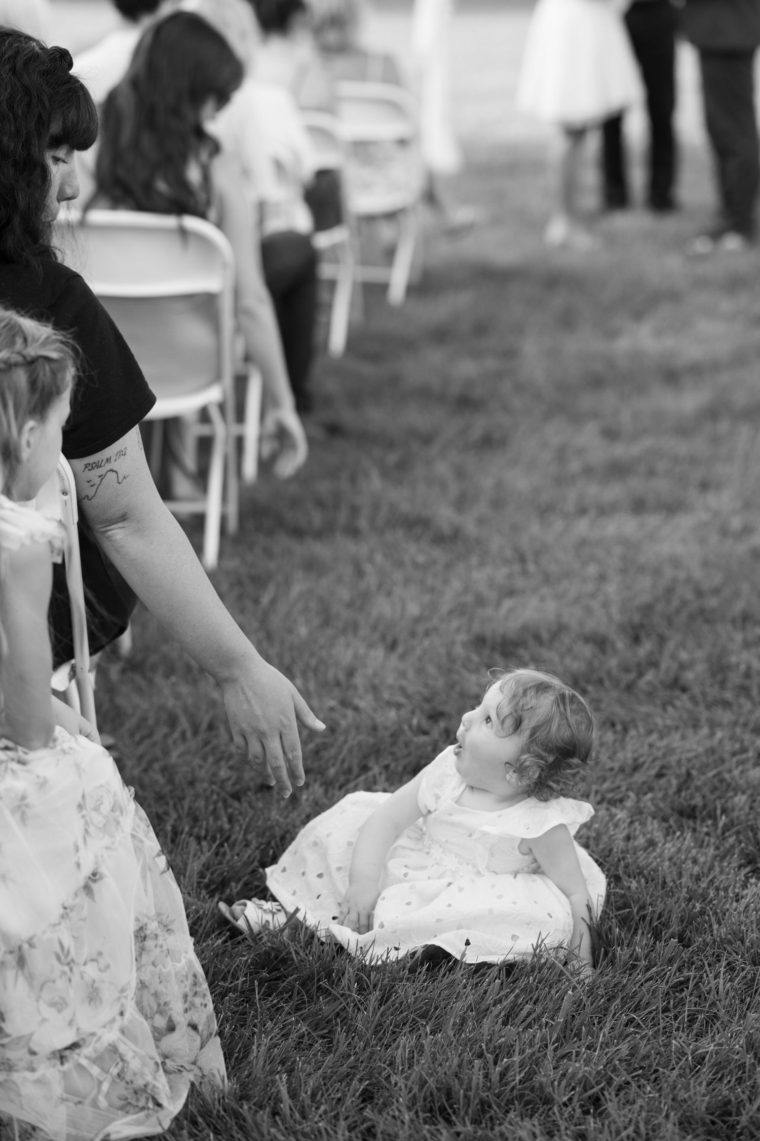 A small toddler in a white dress sits on grass while someone in lace reaches toward them at an outdoor event.