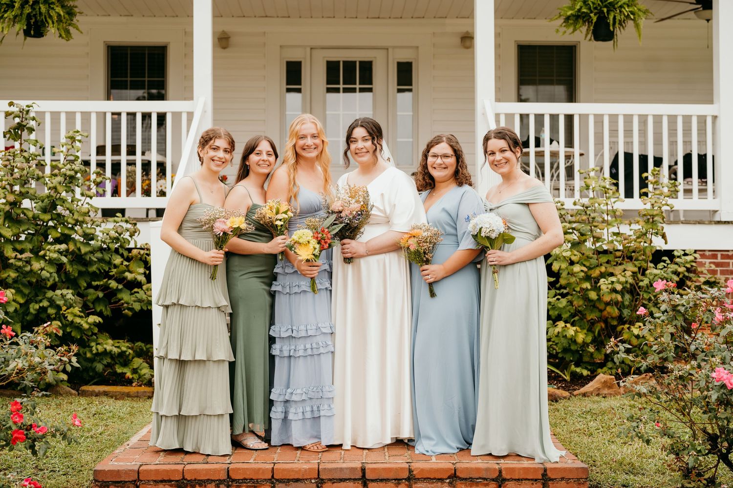 Bridesmaids wearing light blue dresses hold bouquets while standing in a row on a white porch.