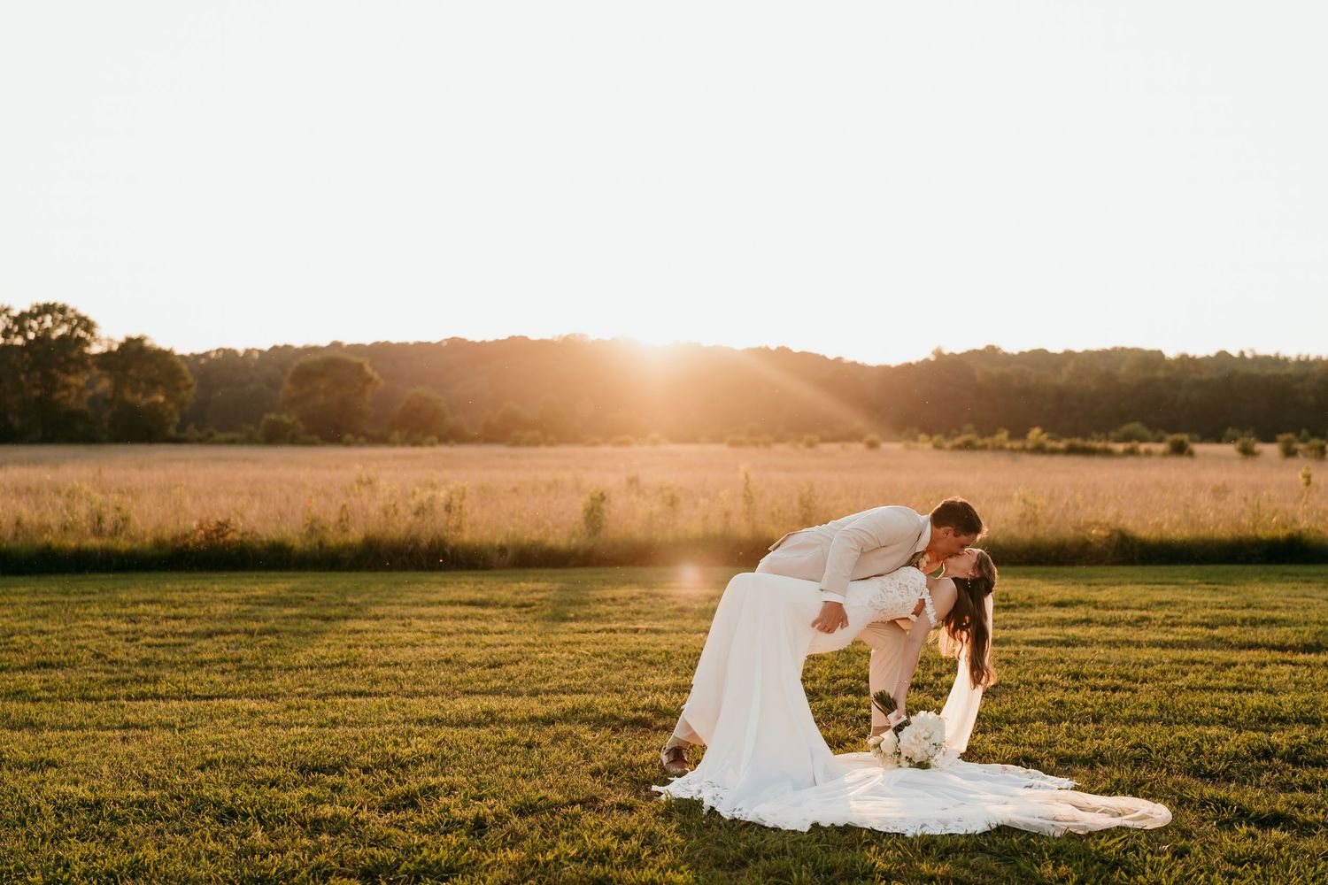 A golden sunset illuminates a grassy field as a couple shares an intimate moment at their wedding.