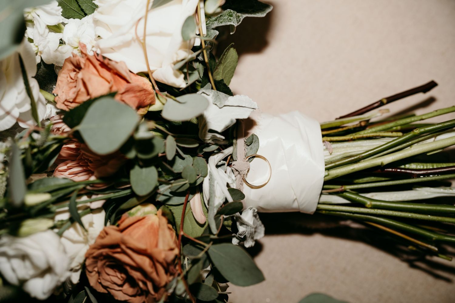 Wedding rings rest on an elegant bouquet of peach roses and white flowers with eucalyptus greenery.