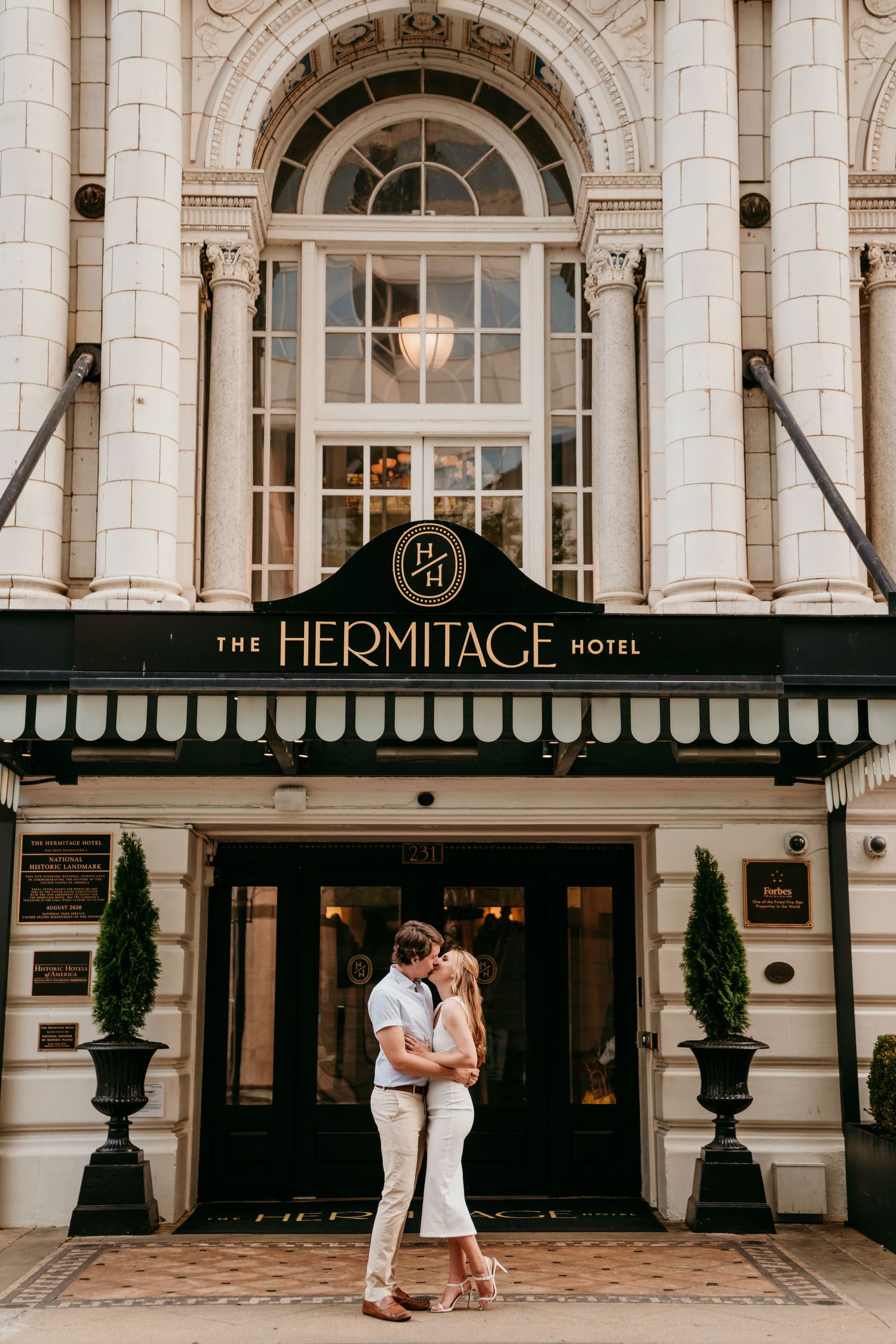 A romantic couple embraces in front of The Hermitage Hotel's elegant stone facade and arched entrance.