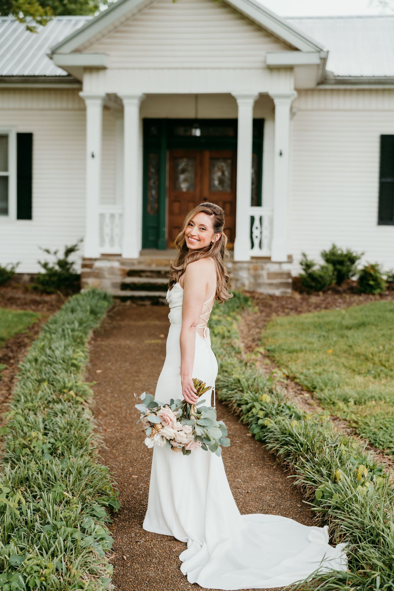 wedding dress stands on a gravel path in front of a classic white farmhouse with columns at battle mountain farm in Nashville TN