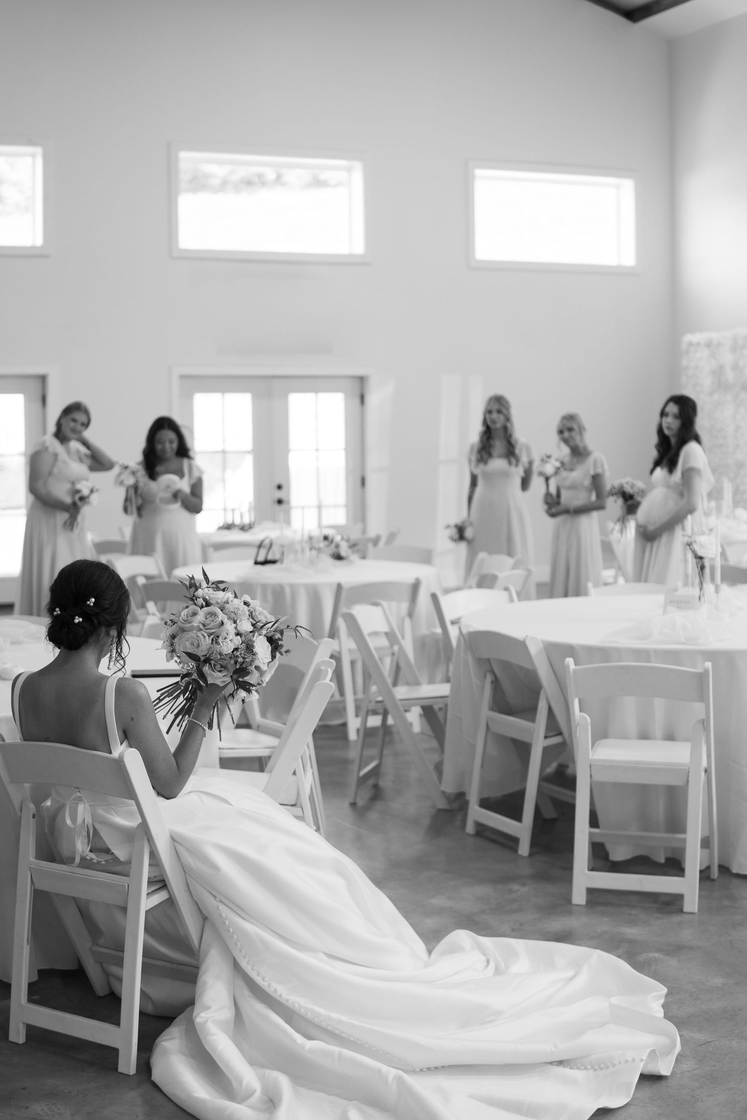 Black and white photo of a wedding reception setup with white chairs and decorations in a bright room with tall windows.
