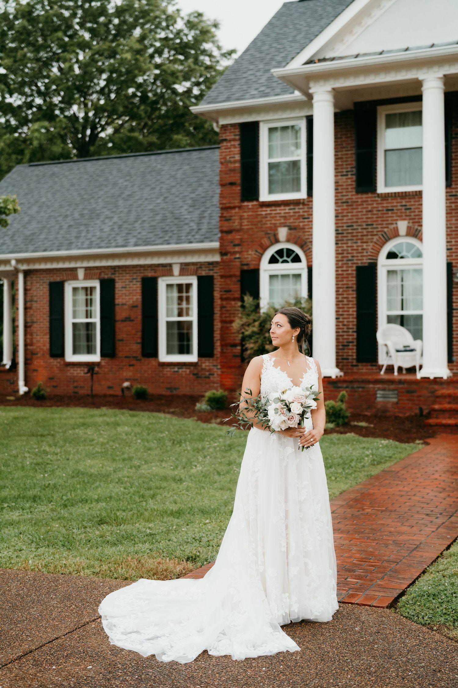 A bride in a flowing white wedding dress poses elegantly on a brick walkway in front of a traditional red brick house.