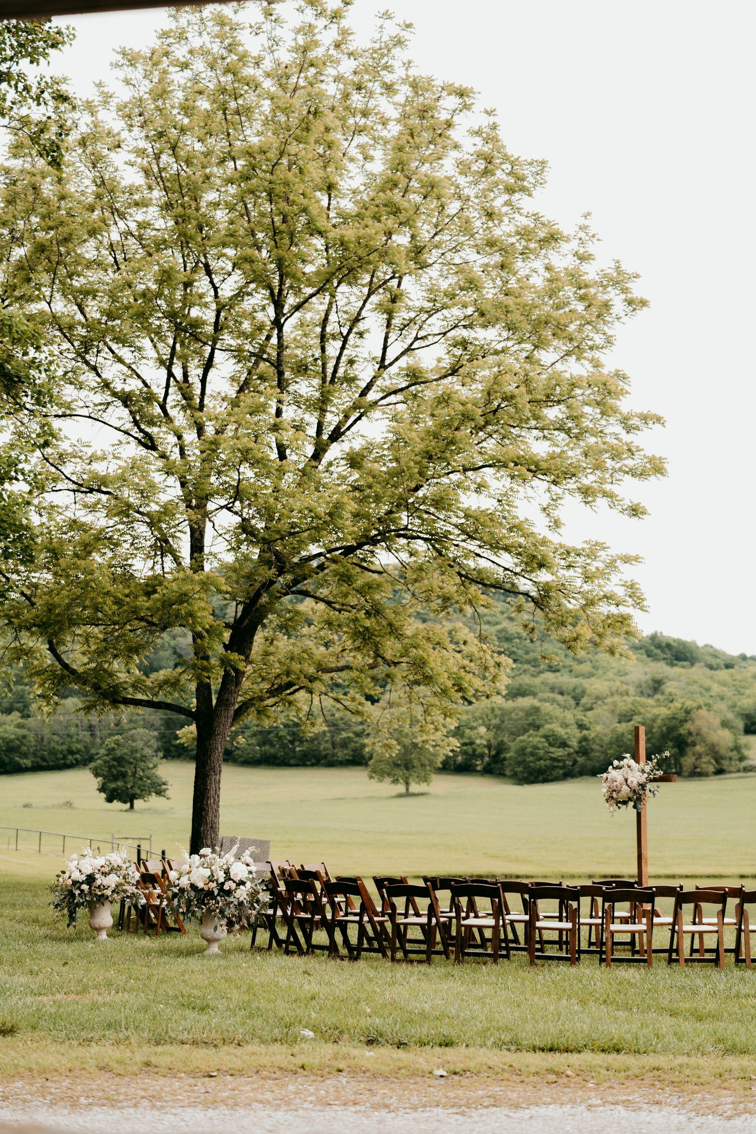 Outdoor wedding ceremony setup with wooden chairs and floral arrangements under a large oak tree in a pastoral field at battle mountain farm