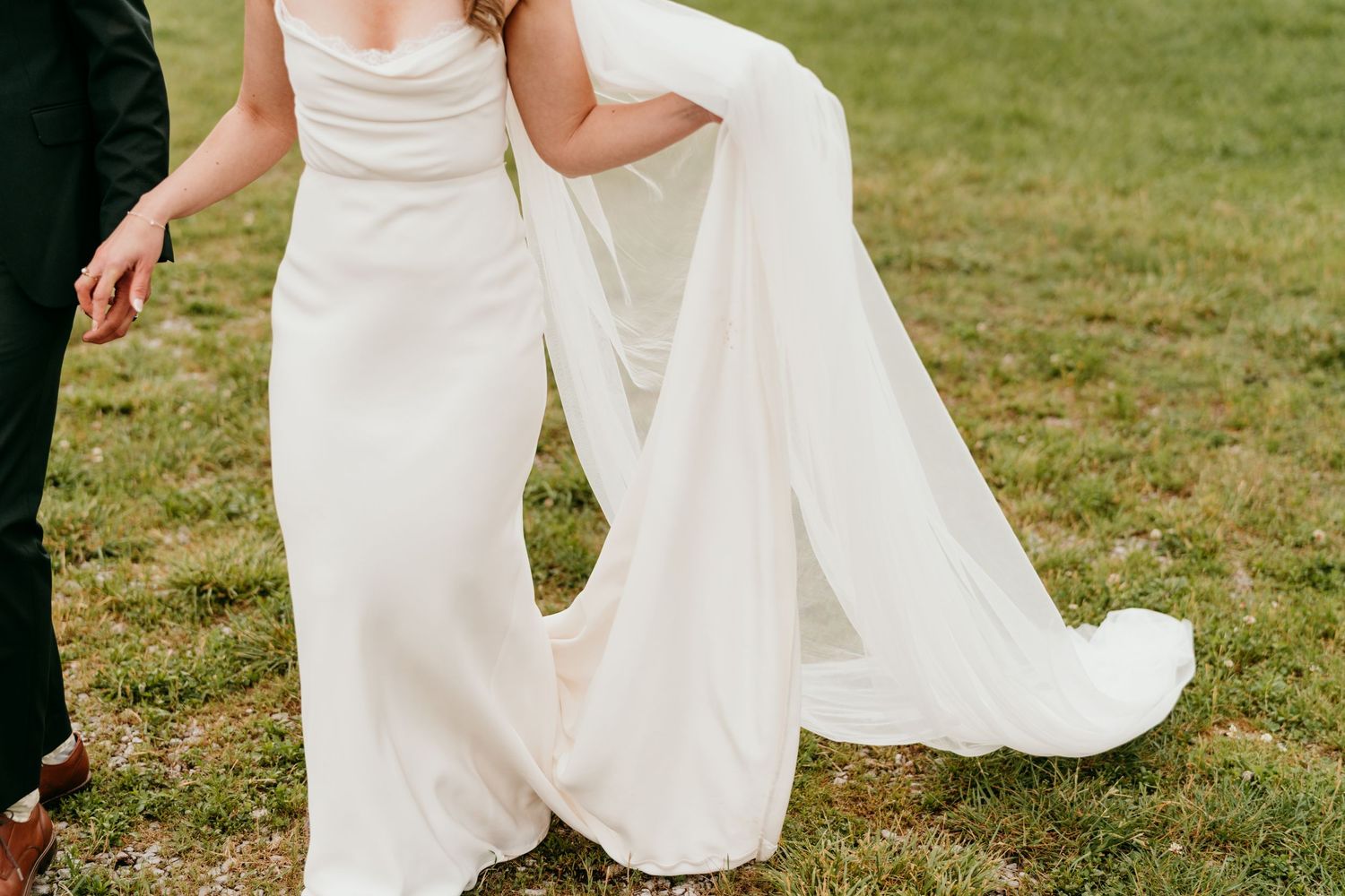 Elegant wedding dress billows in the wind across a sunlit green field.