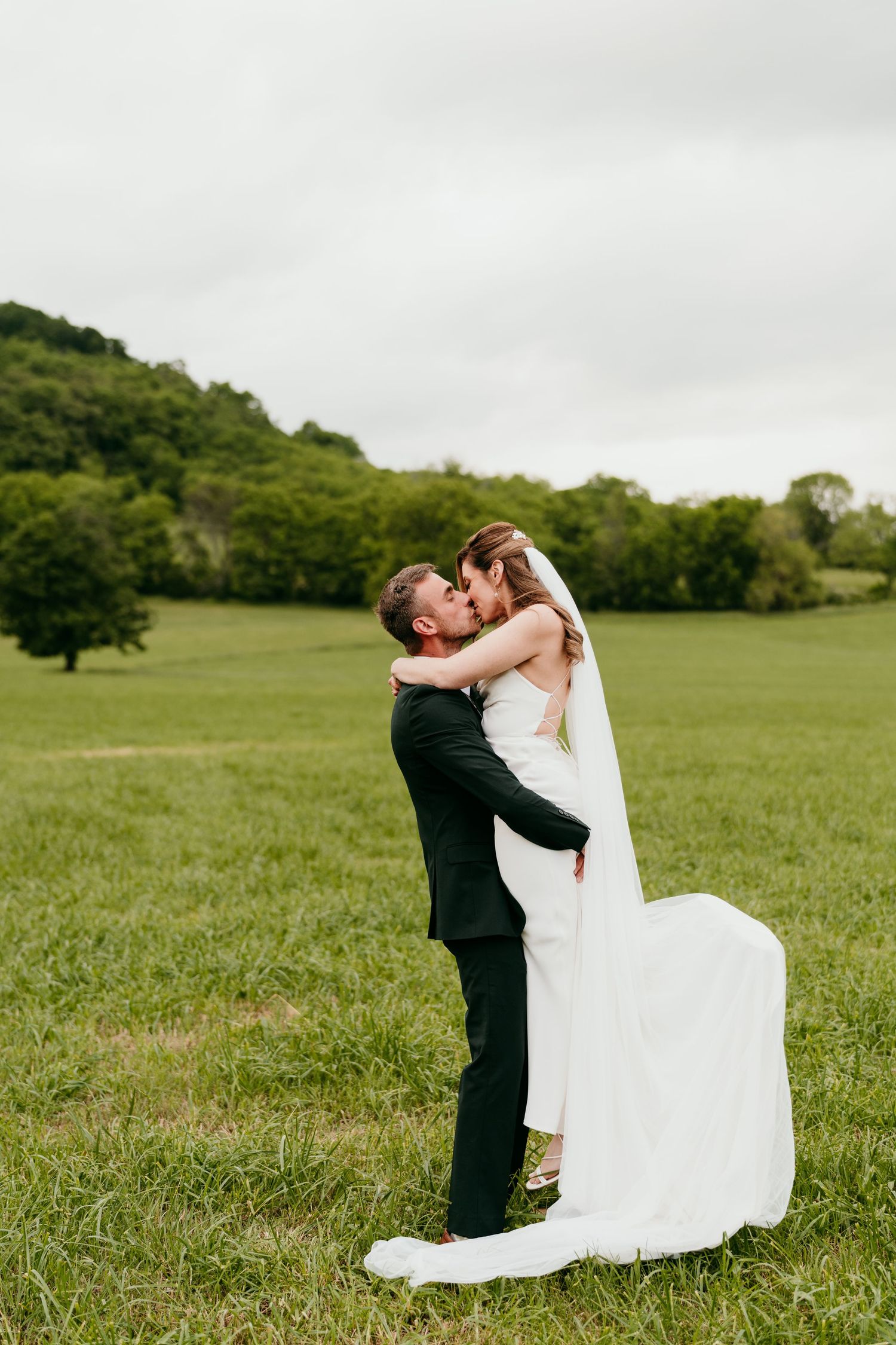 Joyful wedding couple embraces in intimate moment on grassy hillside during outdoor celebration.