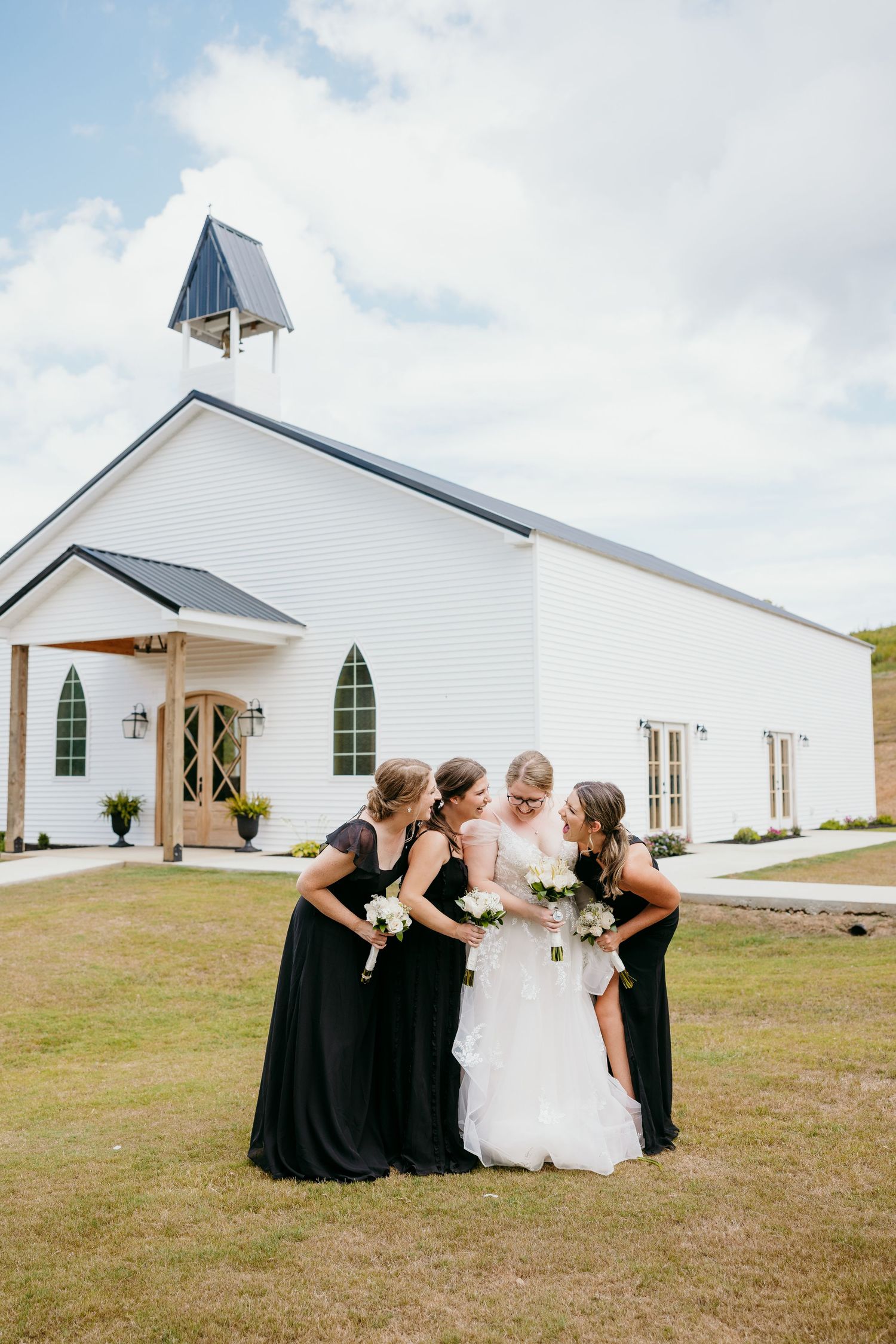 A white rural church wedding venue showcases bridesmaids in black dresses posing on a grassy lawn with the bride.
