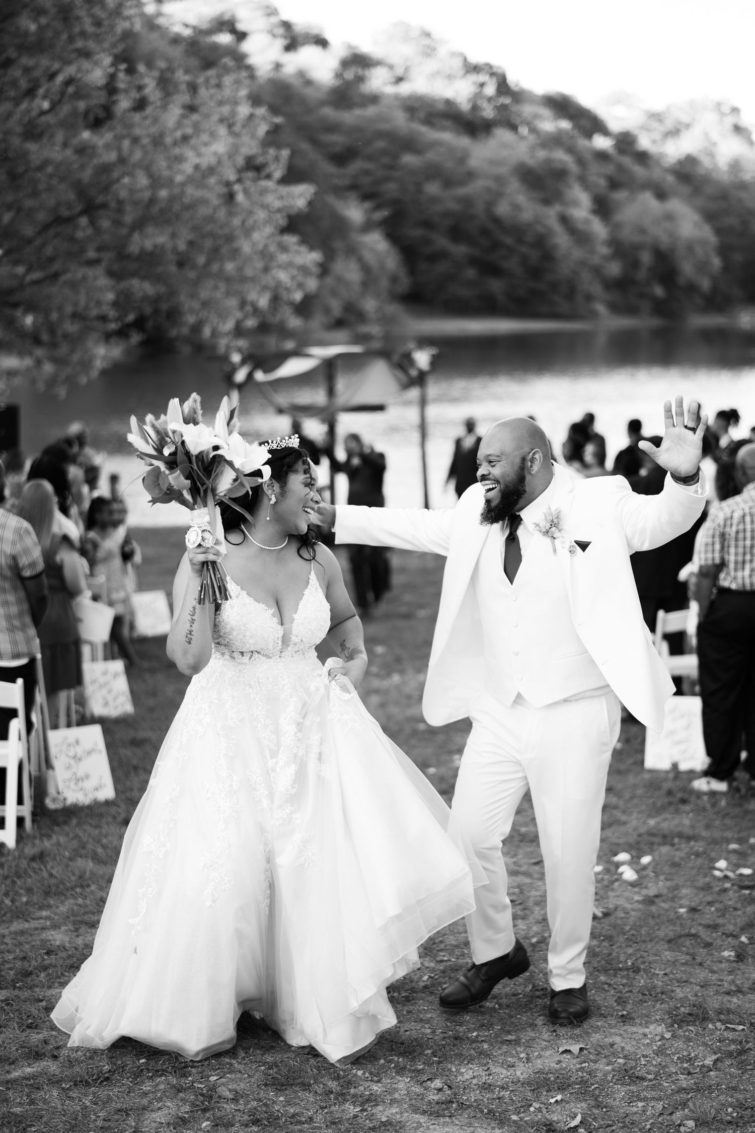 People in white formal attire walk and dance in a joyful procession through an outdoor grassy area during a wedding celebration.