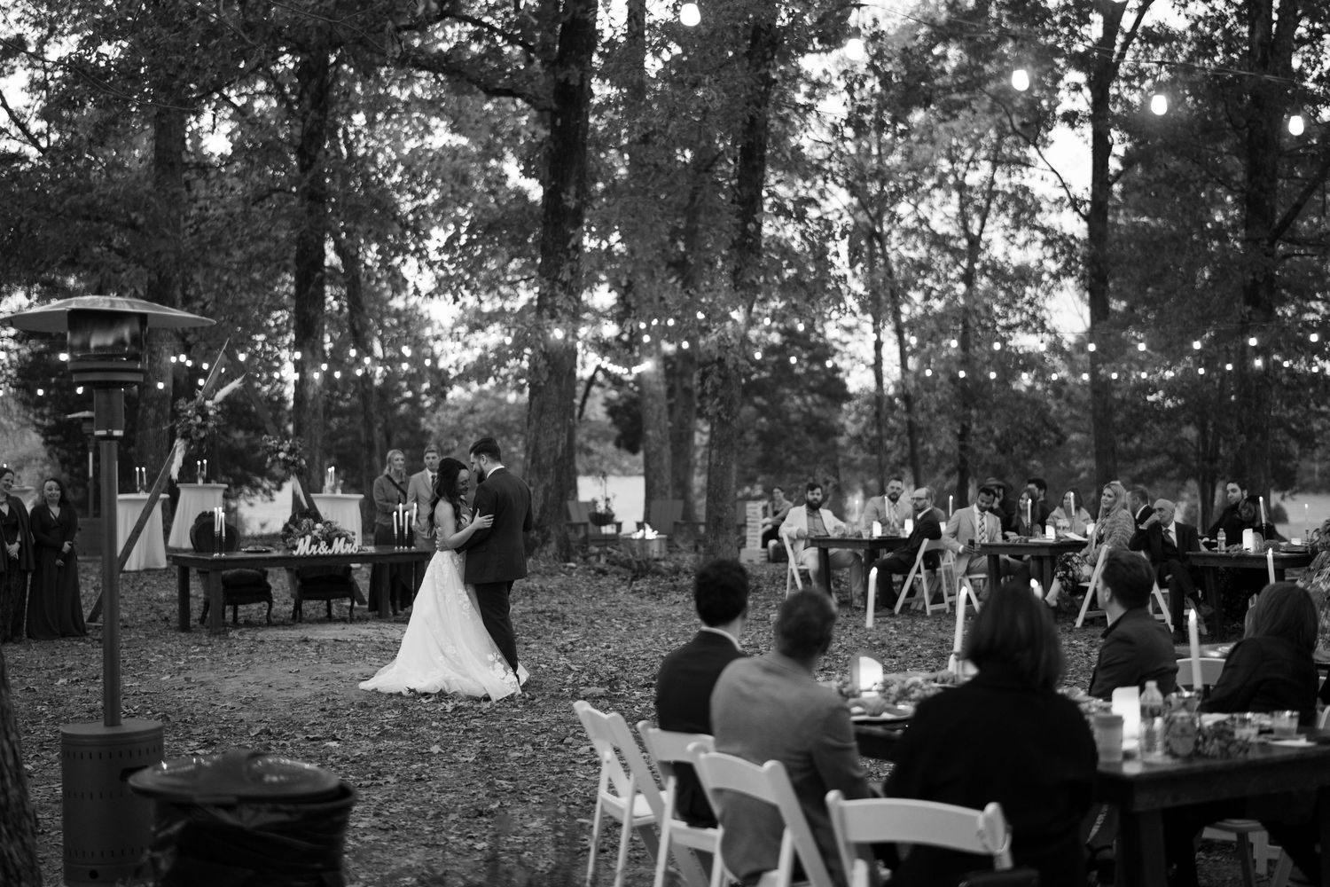 Black and white panoramic view of an outdoor wedding reception under tall trees.
