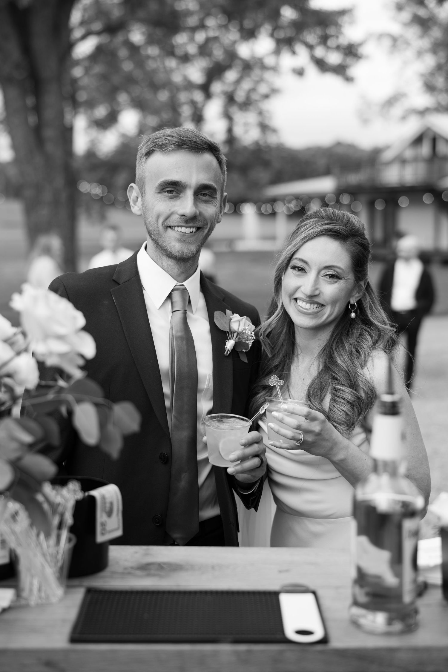 Newlywed couple smiles while holding drinks at outdoor wedding reception bar in black and white photography.