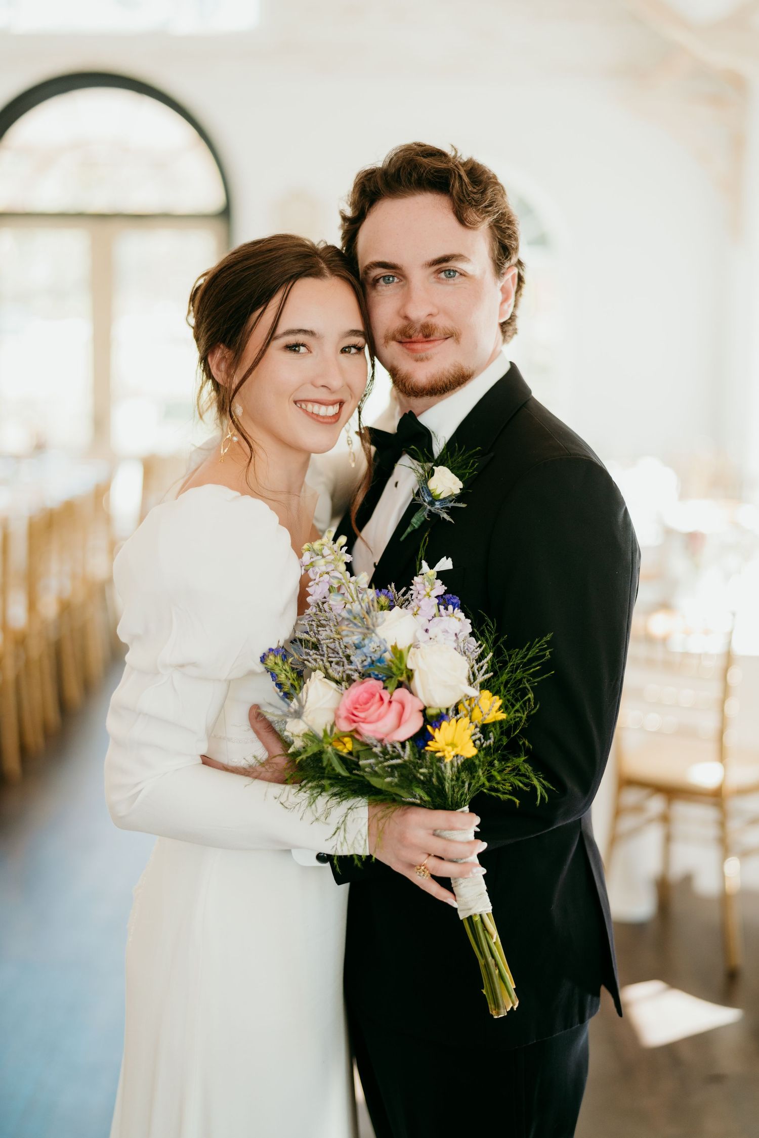 A romantic wedding couple poses together in a bright sunlit setting with colorful floral bouquet.