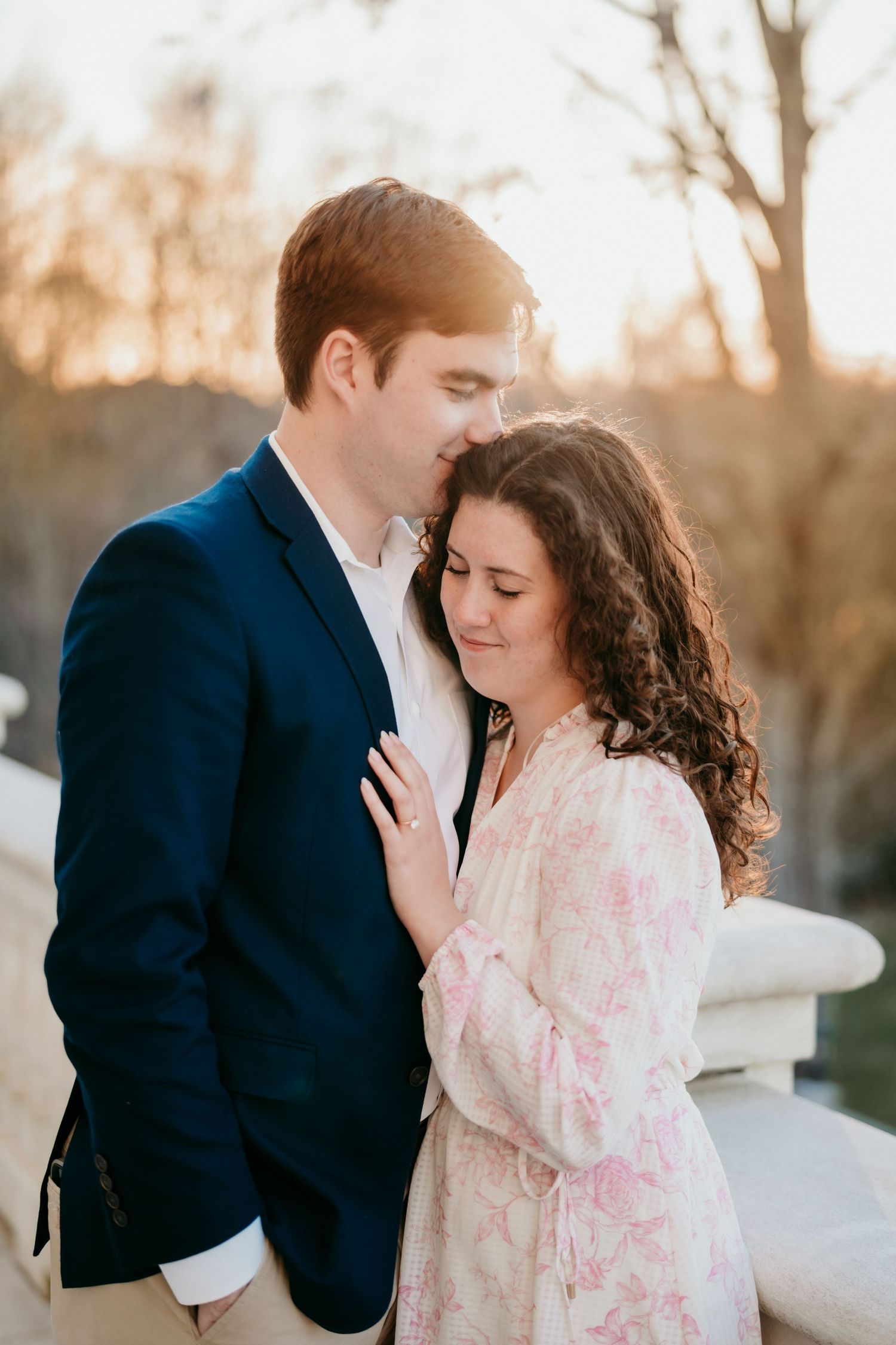 A couple shares an intimate moment at sunset, with her in a floral dress and him wearing a navy blazer.