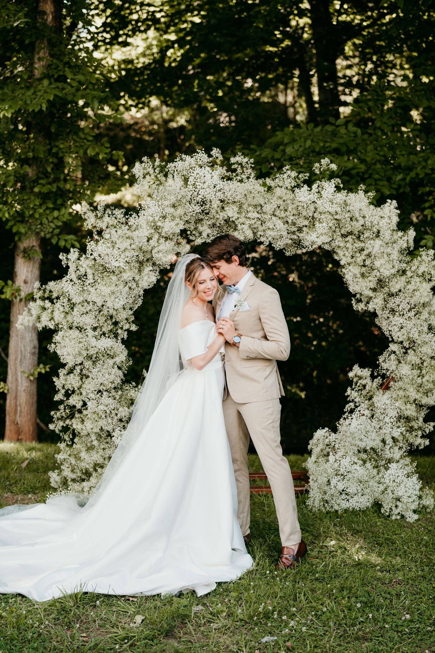 Wedding couple poses under spectacular white floral arch in garden setting.
