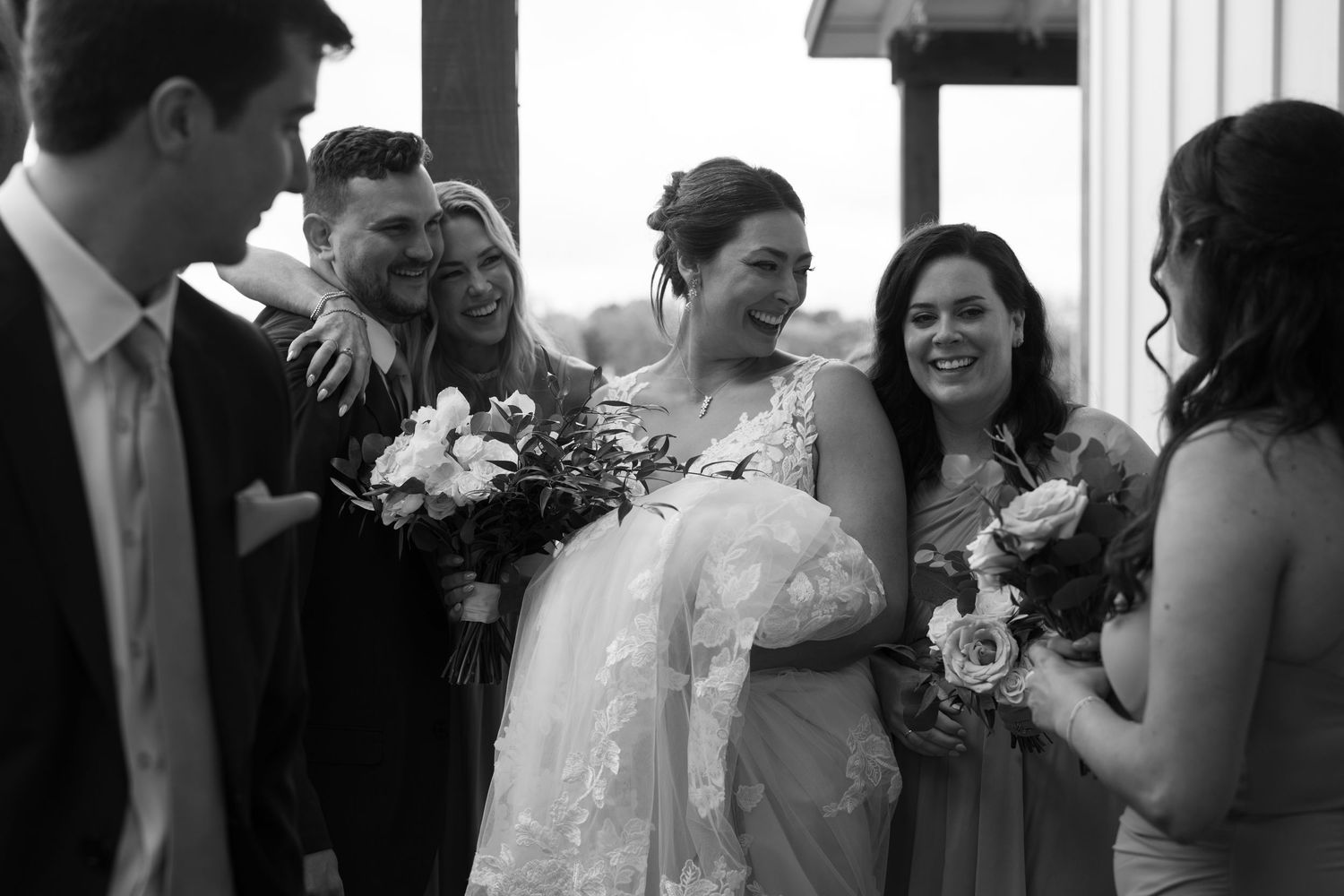 A wedding party laughs together in a candid black and white moment, with the bride in white holding a floral bouquet.