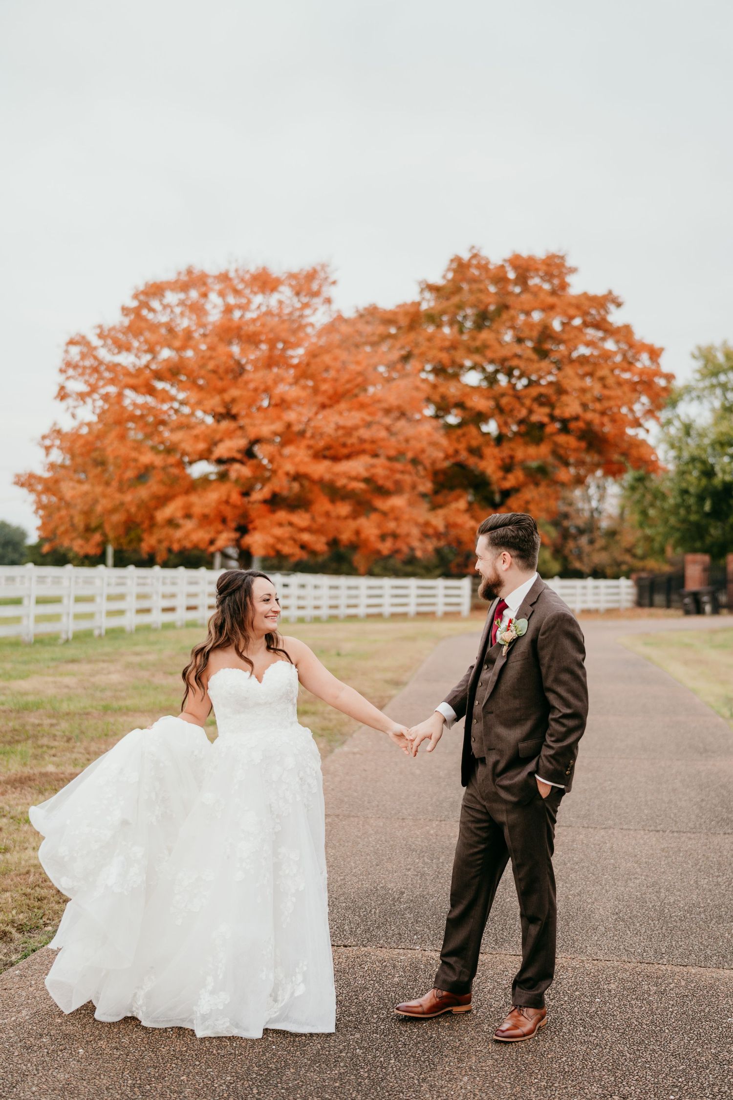 A couple in wedding attire holds hands on a pathway with vibrant autumn trees and white fencing in the background.