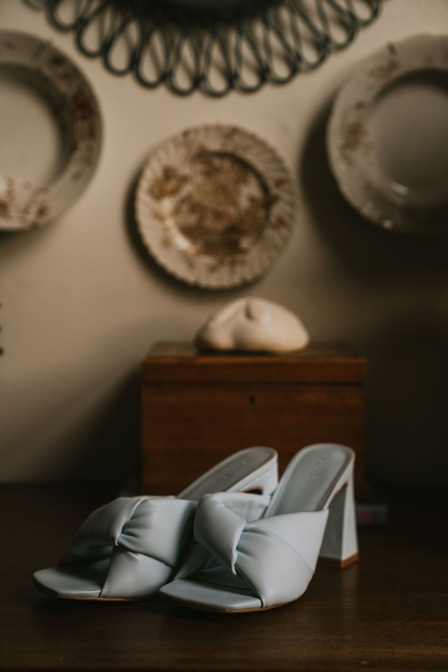 Pale blue wedding shoes sit on a dark wooden surface beneath decorative wall plates.