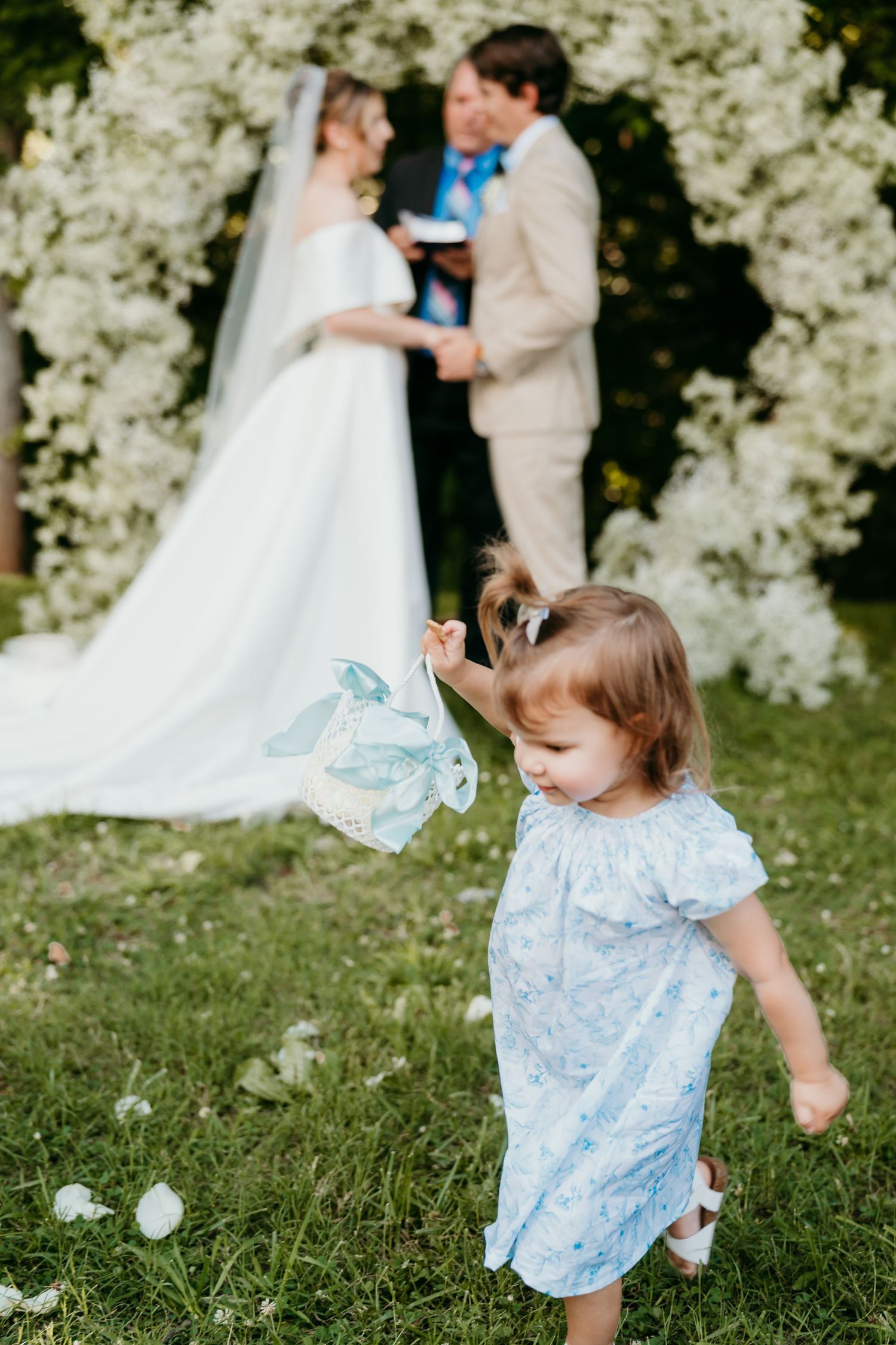 A small child in a blue dress plays with bubbles on a grassy lawn during an outdoor wedding ceremony.