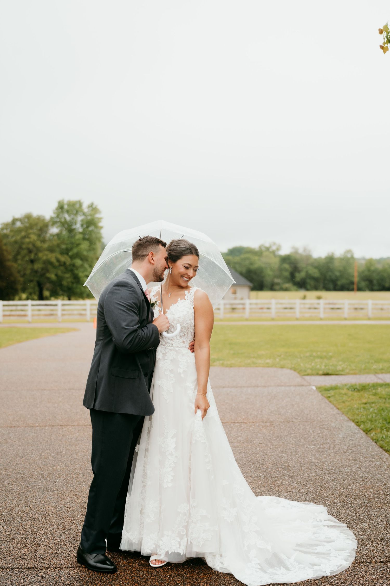 Wedding couple poses together on a gravel path with white fencing in the background.