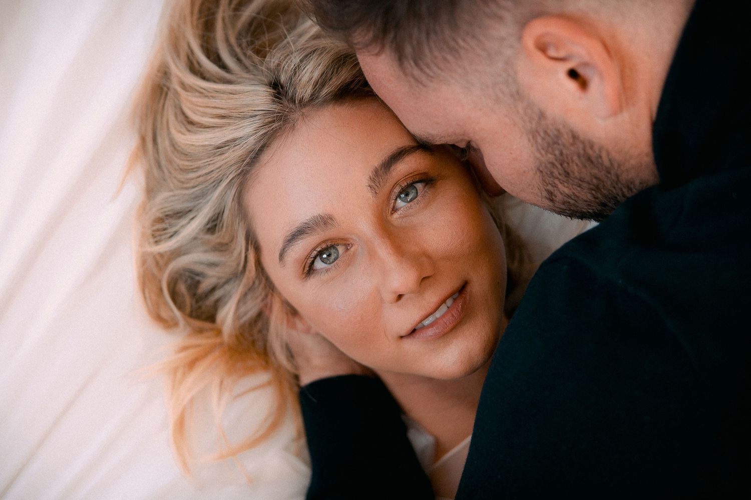 An intimate close-up portrait shows a couple sharing a tender moment against white bedding.
