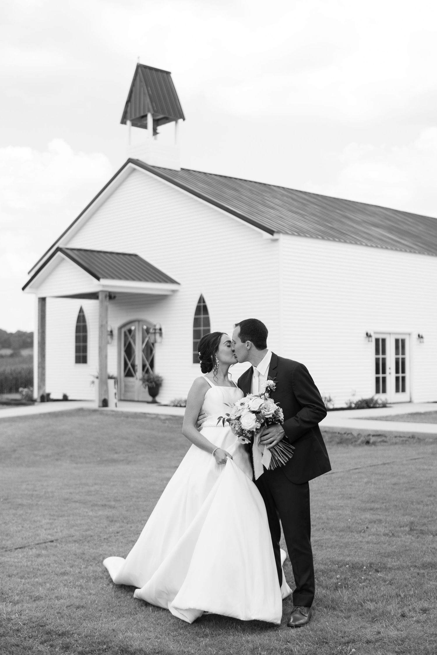 A romantic couple shares a dip dance pose on grass in front of a white chapel during their wedding celebration.