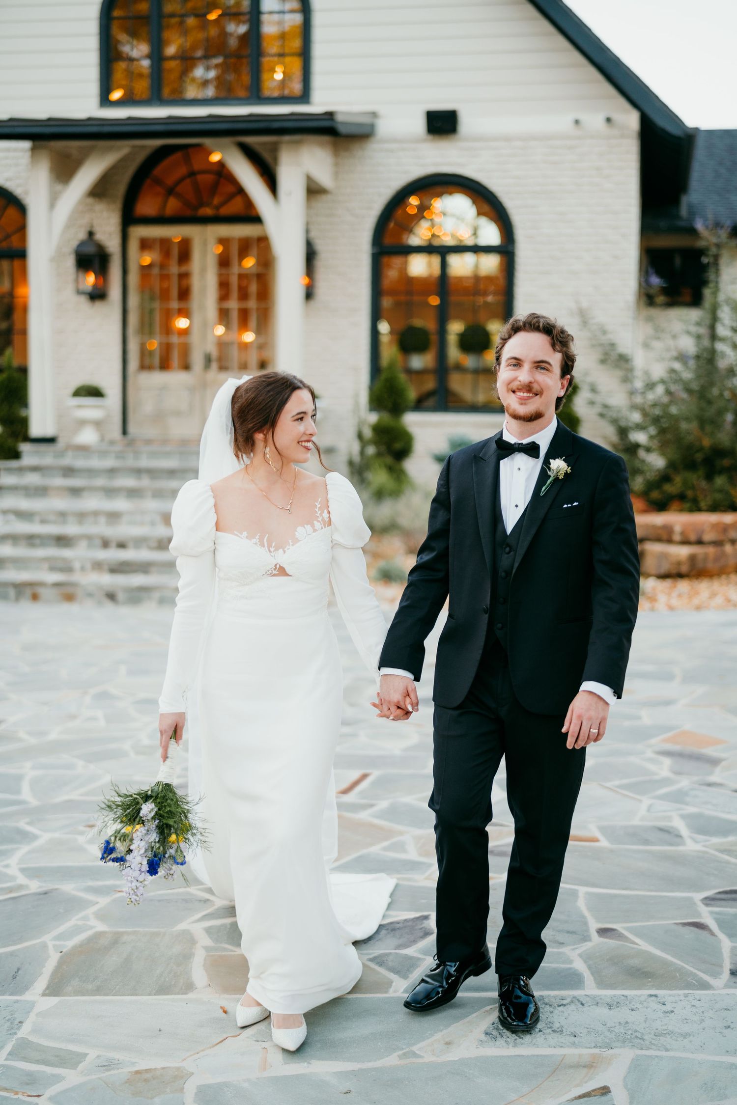 Newlyweds pose together on stone steps outside an elegant white building with arched windows.