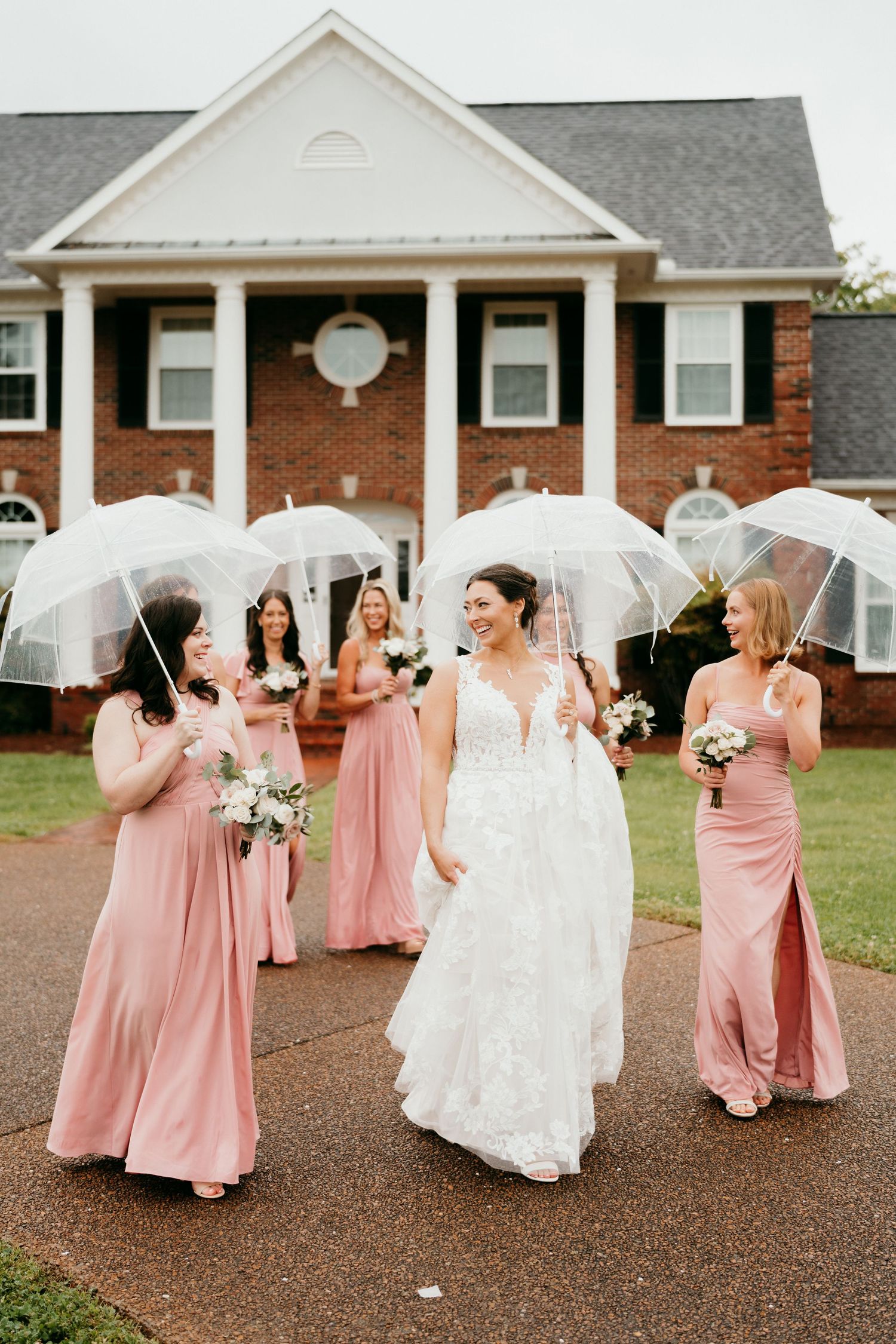 Bridal party in pink dresses holding clear umbrellas walks together on rainy wedding day outside brick building.