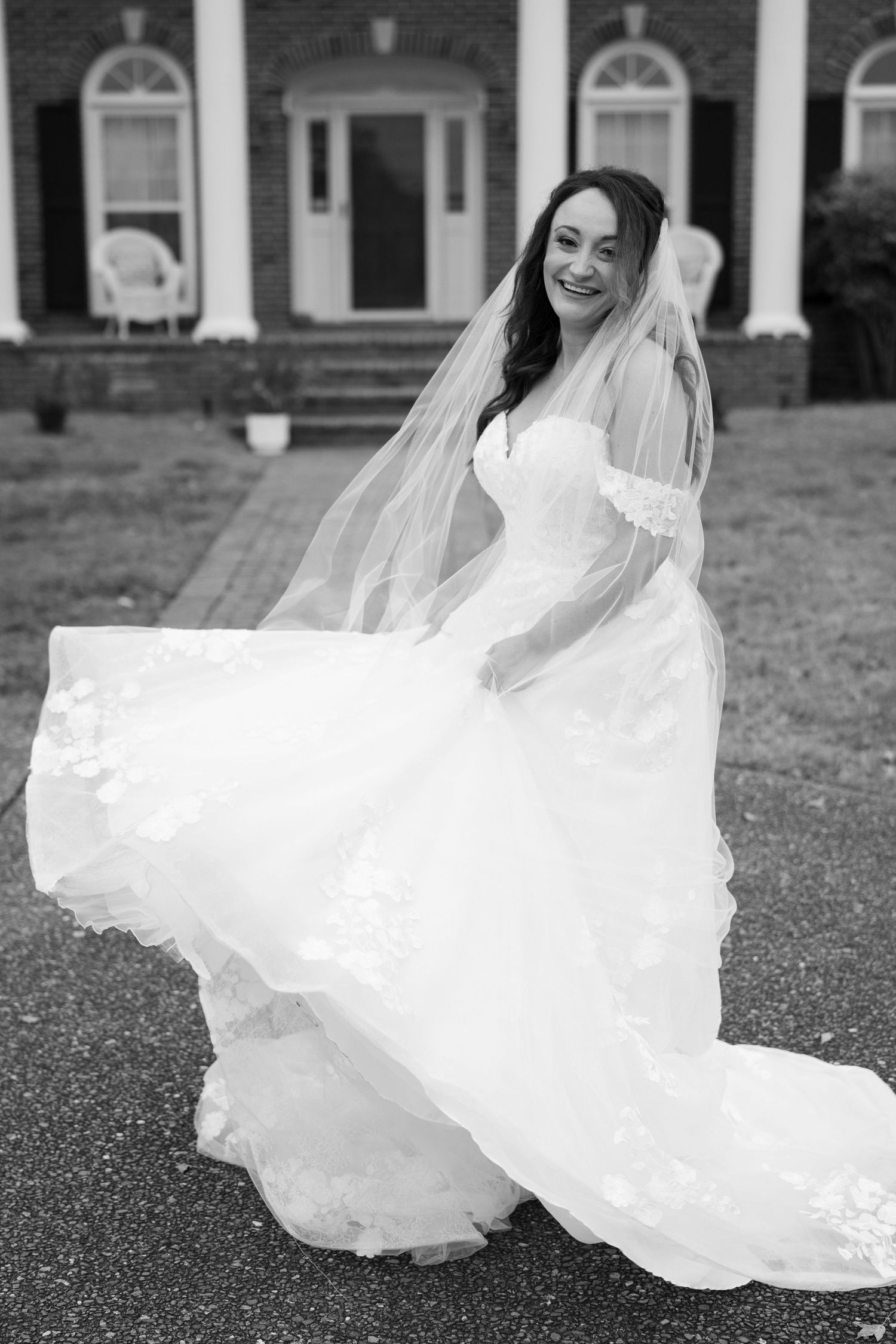 Black and white photo of a joyful bride twirling in her flowing white wedding dress and veil outside a brick building.